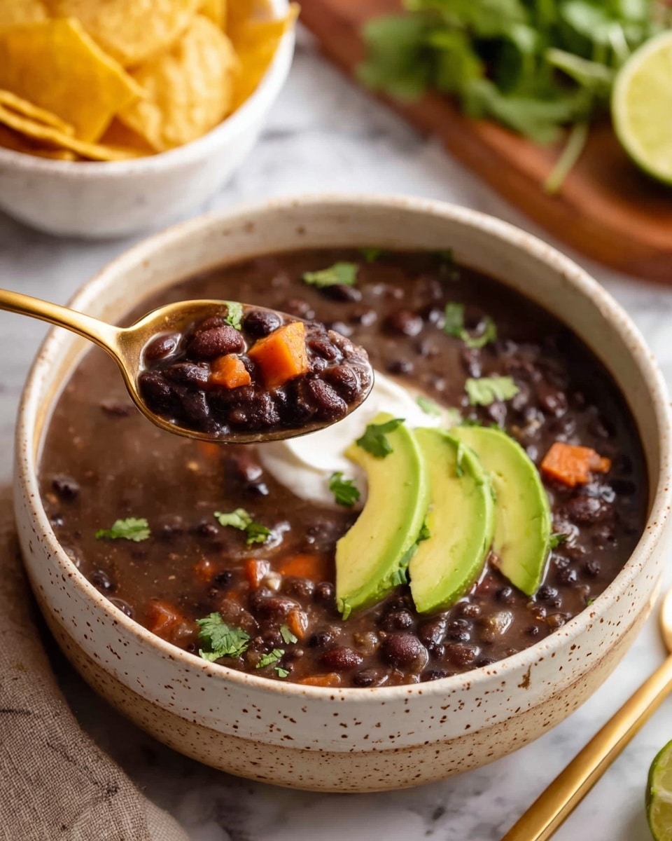 A bowl of black bean soup with visible black beans, small orange carrot pieces, and light brown onion bits in dark brown liquid fills the speckled beige bowl. On top, there is a layer of white sour cream in the center, three pale green avocado slices slightly curved around the sour cream, and several bright green cilantro leaves scattered over the soup. The bowl sits on a white marbled surface next to a white bowl filled with light yellow tortilla chips. In the background, a wooden cutting board holds lime wedges and more green cilantro. A gold spoon lies on a red and white patterned cloth to the left of the soup bowl. Photo taken with an iphone --ar 4:5 --v 7