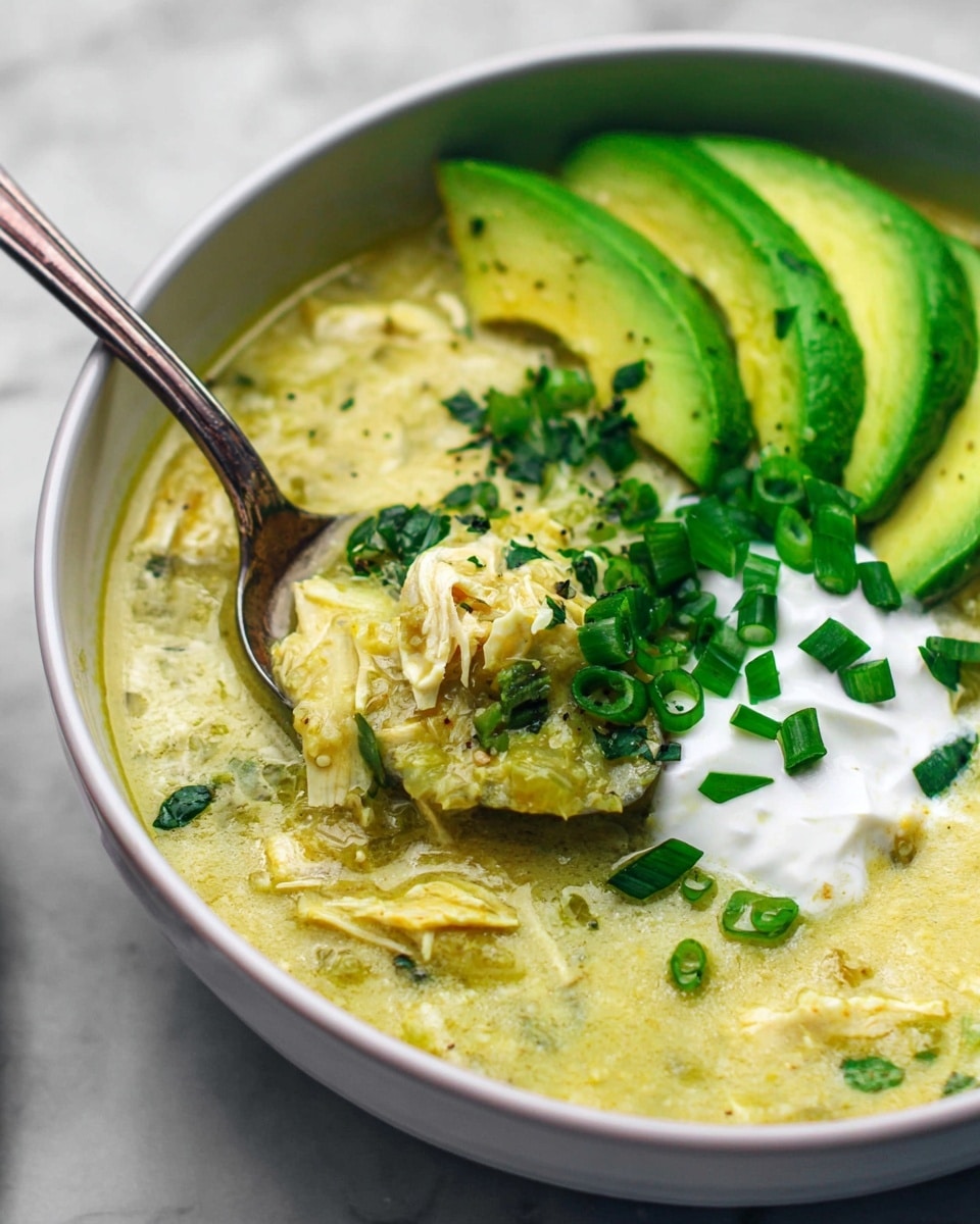 In a white bowl on a white marbled surface, there is a creamy, thick greenish-yellow soup with shredded pieces in it. On the right side of the soup, there are three slices of bright green avocado placed neatly in a row, topped with chopped dark green scallions. In the center, a dollop of white cream is sitting under more scallions. A spoon is scooping some of the thick soup mixed with shredded pieces from the left side of the bowl, showing the creamy texture and bits of herbs. photo taken with an iphone --ar 4:5 --v 7