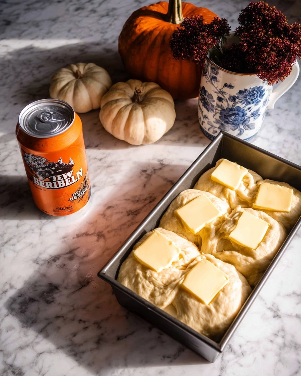A rectangular metal loaf pan on a white marbled surface holds a thick dough layer spread evenly across the base with seven large, pale yellow butter slices placed on top in two rows; to the left, an opened orange soda can with