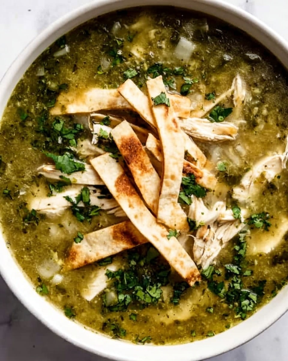 The image shows a white bowl filled with a green salsa soup, topped with shredded light beige chicken pieces scattered around the surface. On one side of the bowl, there are several strips of toasted tortilla placed on top of the soup, their color a mix of light brown and golden with some darker grill marks. The soup has a slightly thick texture with visible bits of green herbs mixed throughout. Chopped bright green cilantro is sprinkled generously over the top, adding a fresh color contrast. The bowl is placed on a white marbled surface. photo taken with an iphone --ar 4:5 --v 7