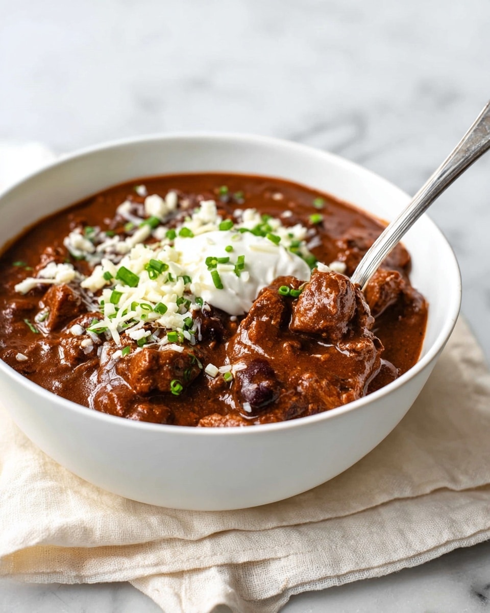 A white bowl filled with thick, dark brown chili stew that has visible chunks of meat throughout, topped with a dollop of white sour cream, small green chopped scallions, and some shredded white cheese scattered on top. A silver spoon is placed in the bowl, partially scooping the chili. The bowl rests on a light cream cloth on a white marbled surface, with a light, blurred background. photo taken with an iphone --ar 4:5 --v 7