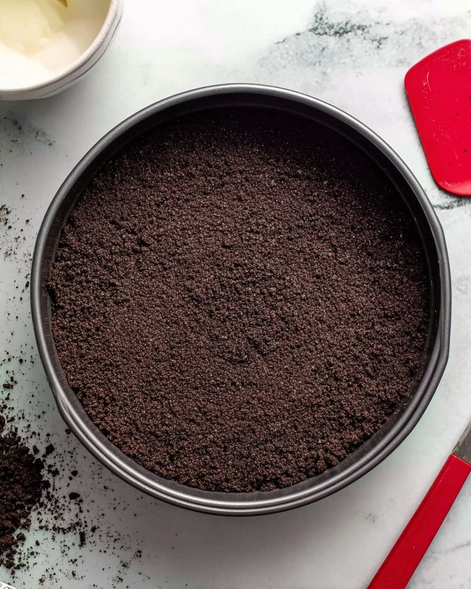 The image shows a single dark brown crumbly layer pressed evenly into a round black springform pan, forming a smooth flat base with slightly raised edges all around. The pan sits on a white marbled surface with scattered black crumbs nearby. A red spatula lies to the right side of the pan, and part of a white bowl with some white creamy substance is visible at the top left corner. photo taken with an iphone --ar 4:5 --v 7