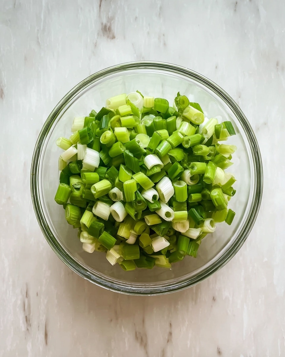 A clear glass bowl sits on a white marbled surface, filled with many pieces of chopped green onions. The green onions have light green and white rings that look fresh and bright, with the pieces stacked loosely in the bowl. The image is shot from above, showing the bowl's round shape and the onions inside clearly. photo taken with an iphone --ar 4:5 --v 7