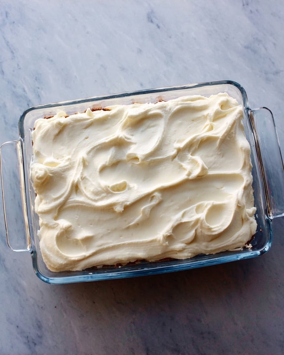 A clear glass rectangular dish with two handles holds a layered dessert. The top layer is thick, smooth, and creamy, whipped with gentle swirls and light peaks, covering the entire surface. Underneath, slightly visible through the dish edges, are darker layers of cake or filling. The dish is set on a white marbled surface. photo taken with an iphone --ar 4:5 --v 7