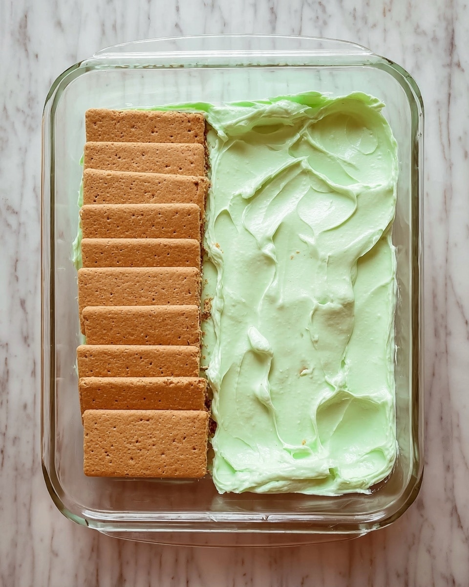 The image shows a glass rectangular dish placed on a white marbled surface. Inside the clear dish, there are two layers visible. The first layer consists of a row of rectangular light brown graham crackers neatly placed side by side at the bottom of the dish. On top of the graham crackers, there is a thick layer of light green creamy mixture with some small darker specks spread unevenly over one half of the crackers. The creamy layer looks smooth with soft peaks and folds, and the scene is shown from above in two different stages of spreading the creamy mixture. photo taken with an iphone --ar 4:5 --v 7