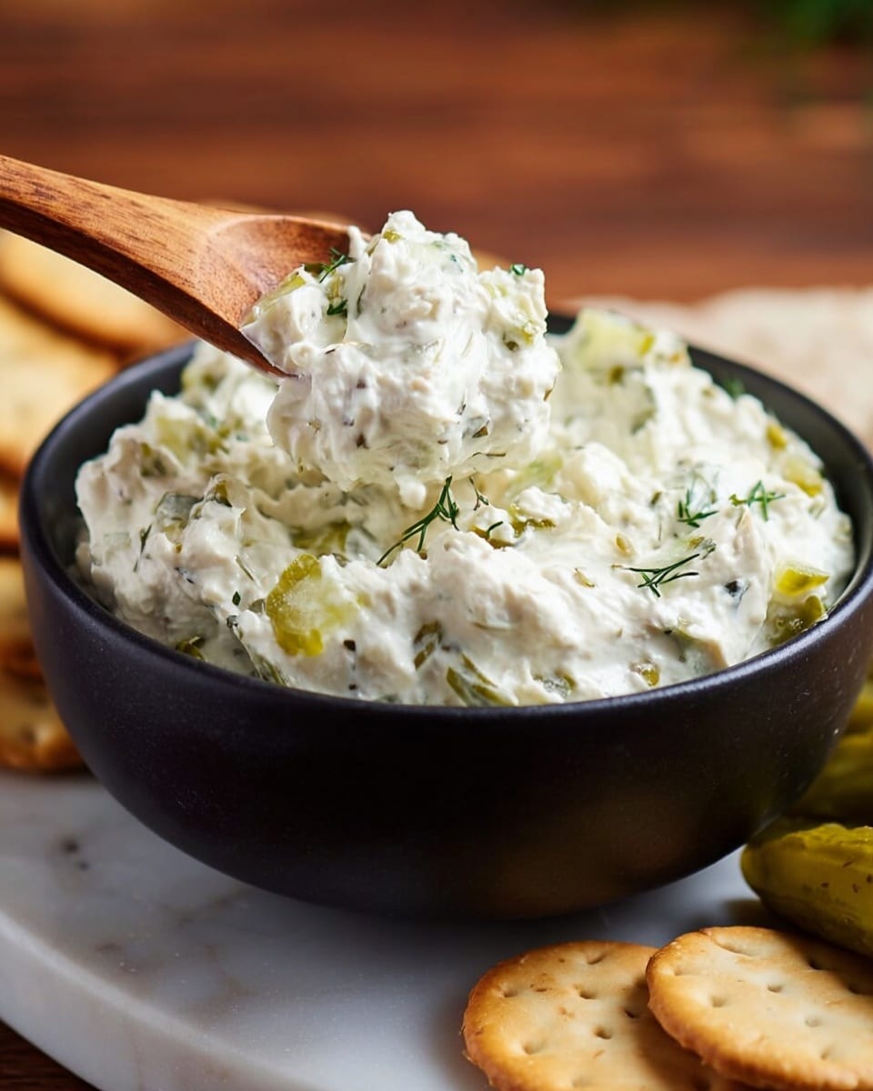 A close-up of a dark bowl filled with creamy white spread mixed with small green pickle pieces and herbs, showing a thick and chunky texture. A wooden spoon lifts some of the spread from the bowl, highlighting its soft and rich consistency. Around the bowl, light brown round crackers and pickle slices are partially visible resting on a white marbled surface, suggesting a ready-to-eat snack. The background is softly blurred with warm wooden tones. Photo taken with an iphone --ar 4:5 --v 7
