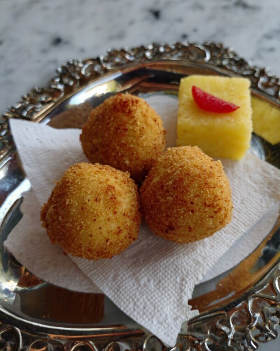 The image shows three round, golden-brown fried balls with a rough texture on a white paper doily placed on a shiny metal tray with intricate cut-out patterns. Next to the fried balls is a small yellow cube with a red piece on top, possibly a garnish or condiment. The background surface is a white marbled texture. photo taken with an iphone --ar 4:5 --v 7