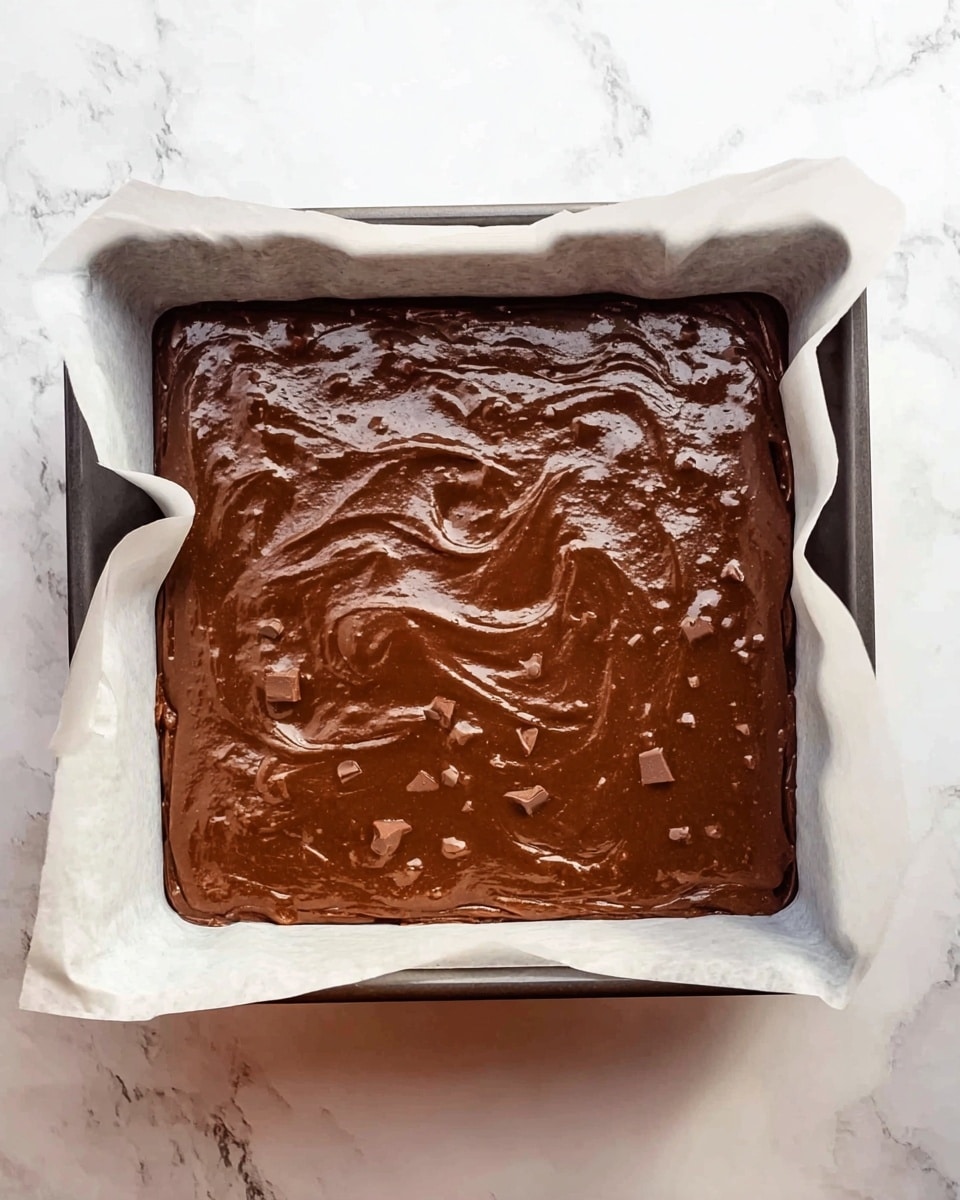 A square baking pan lined with white parchment paper holds a thick layer of smooth, dark brown chocolate batter mixed with small chunks that add texture. The batter is evenly spread to the edges, showing some swirls and ripples on the surface. The pan is placed on a white marbled surface, giving a clean and bright background. Photo taken with an iphone --ar 4:5 --v 7