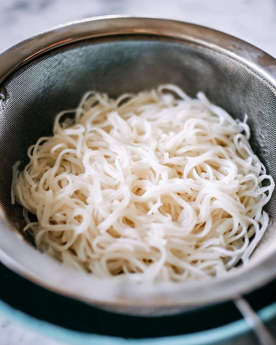 A close-up image of white flat noodles sitting in a round metal strainer above a bowl. The noodles are slightly shiny and soft, tangled loosely in the strainer's mesh. The metal strainer has a silver rim and a fine net texture, resting on a bowl with a light blue rim. The background is blurred but is a white marbled texture. Photo taken with an iphone --ar 4:5 --v 7