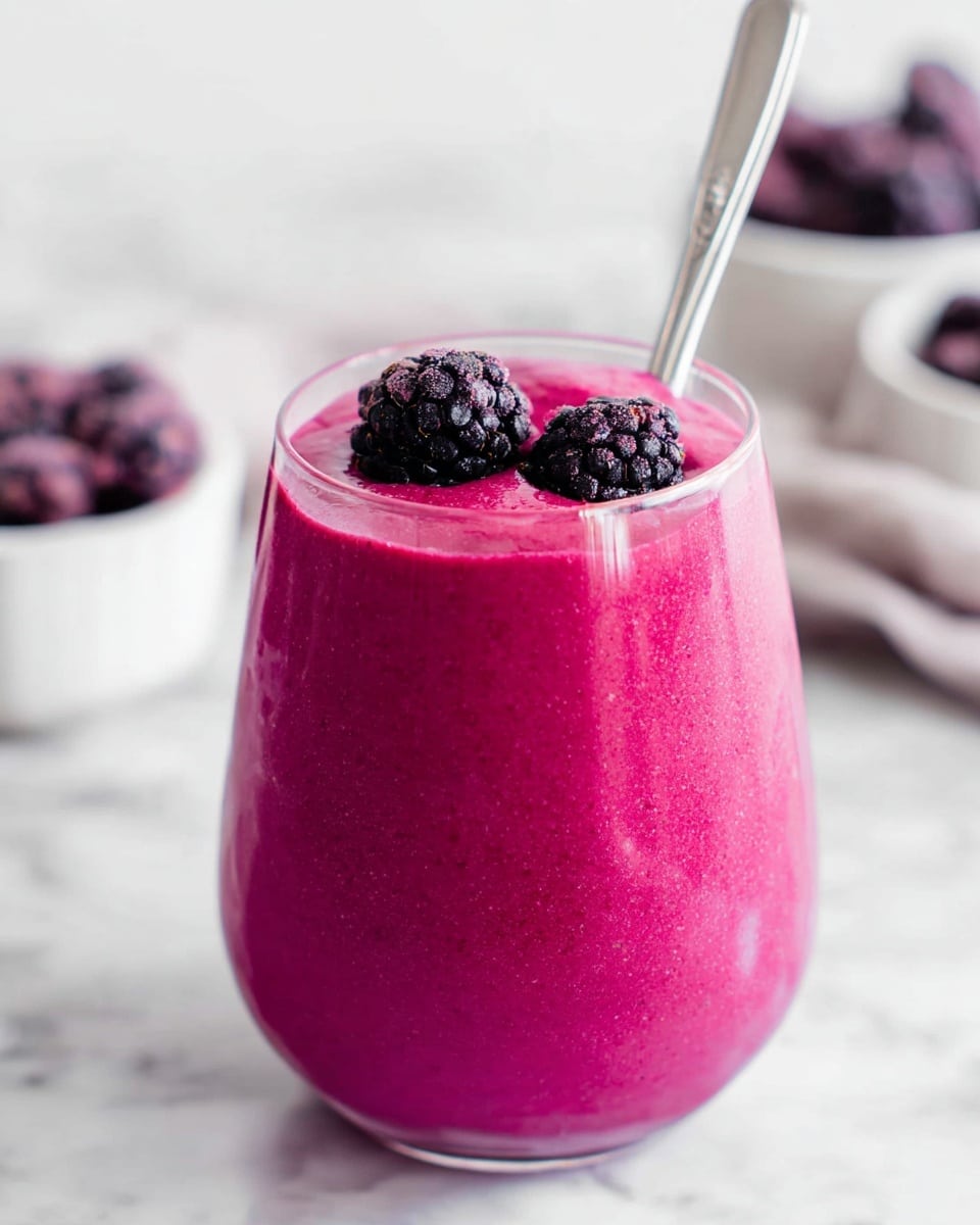 A clear glass filled with two layers of thick, bright magenta smoothie with a smooth texture. On top, there are two dark purple blackberries sitting close to each other. A silver spoon is placed inside the glass, leaning slightly to the left. The glass sits on a white marbled surface. In the blurry background, some white bowls contain similar dark purple berries. photo taken with an iphone --ar 4:5 --v 7