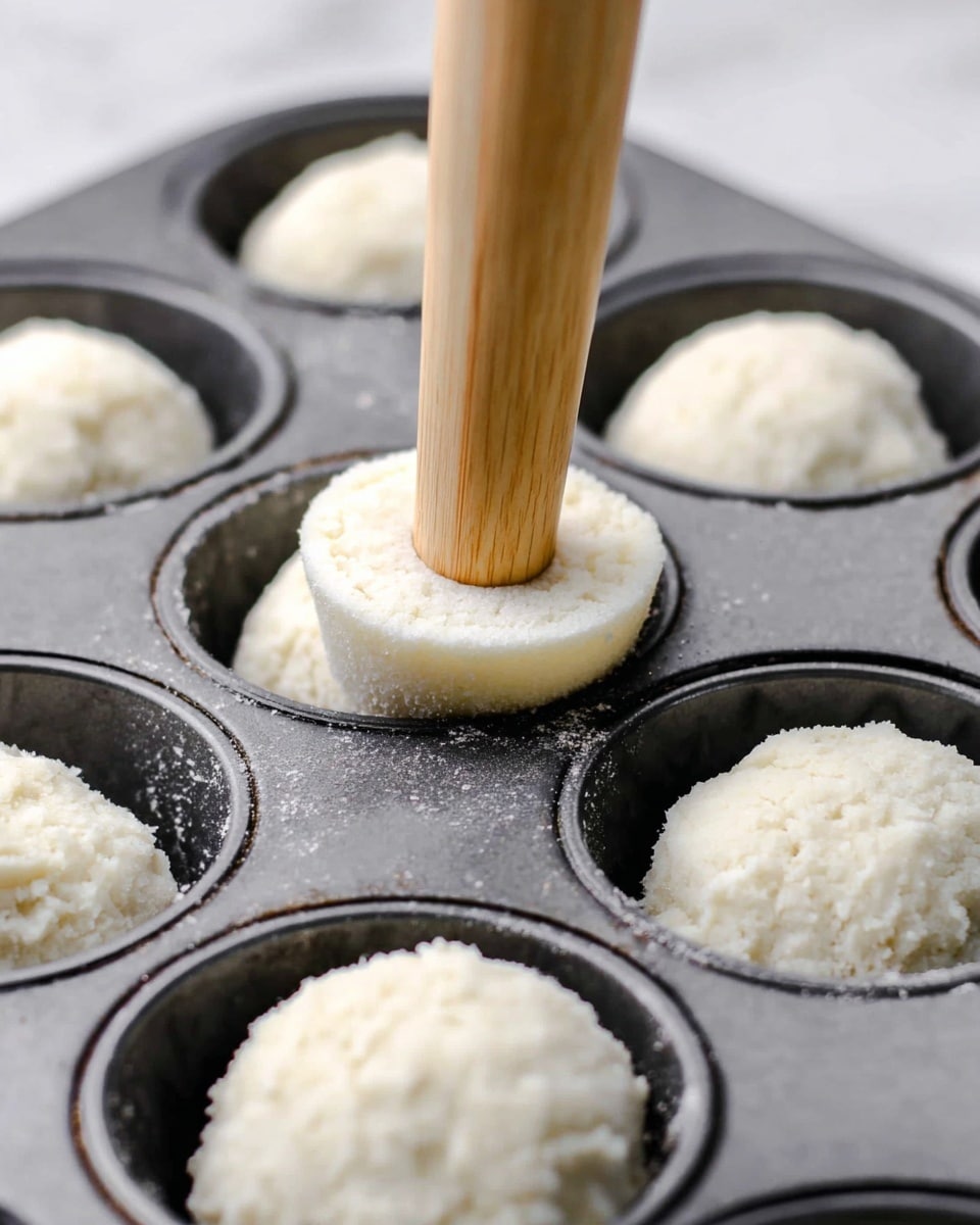 A close-up image shows a dark gray muffin pan with its cups filled with small, round, white dough balls and one circle of white dough pressed into the side of a cup by a wooden tamper with a light natural wood texture. The dough looks soft and fluffy with a light, slightly rough texture, sitting inside the smooth cups of the pan resting on a white marbled surface. photo taken with an iphone --ar 4:5 --v 7