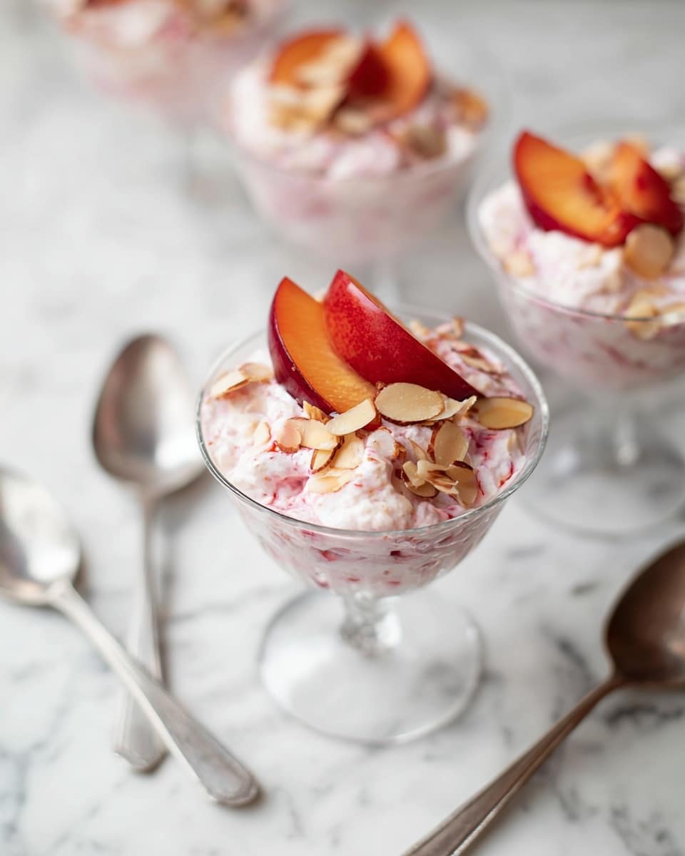 The image shows three clear stemmed glasses filled with a creamy pink dessert that has a chunky texture, likely from fruit mixed in. Each glass is topped with thin slices of peach and some toasted nuts, which add a light brown touch. The glasses sit on a white marbled surface with two small silver spoons nearby. In the background, there is a blurred white plate with dark red plums and a wooden chair with a light-colored cloth draped over it. The lighting is soft and natural, creating a fresh and inviting look. photo taken with an iphone --ar 4:5 --v 7
