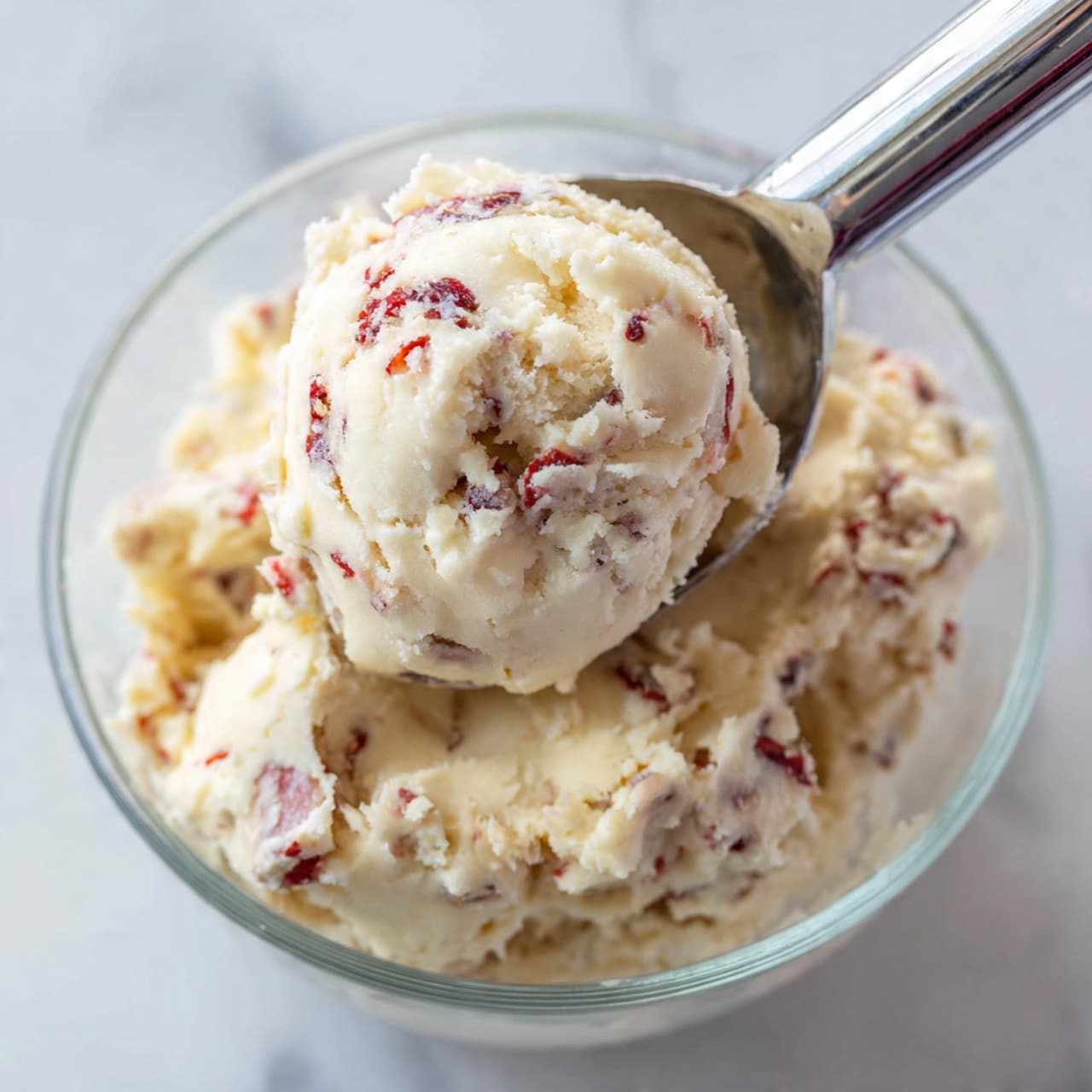 A close-up view of round cookies piled on a white plate, each cookie coated in a thick layer of white powdered sugar with small red specks scattered across the surface. The cookies have a textured, slightly crumbly look underneath the sugar, showing a soft beige color. The white powdered sugar is unevenly spread, creating a rough, snowy effect. The white plate is set against a white marbled background, enhancing the light and soft tones of the cookies. photo taken with an iphone --ar 4:5 --v 7