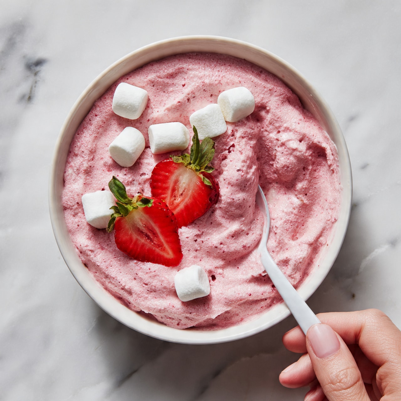 The image shows a white bowl filled with a thick, creamy pink mixture that has a textured, slightly lumpy surface. Scattered over the top are small white marshmallows, adding a soft, pillowy contrast to the pink base. Near the center, two bright red strawberry halves with green leaves sit upright, adding a fresh, colorful touch. A spoon with a white handle rests partially inside the bowl, with a woman's hand holding it from the bottom right. The bowl is placed on a white marbled surface that gives a clean, elegant background. Photo taken with an iphone --ar 4:5 --v 7