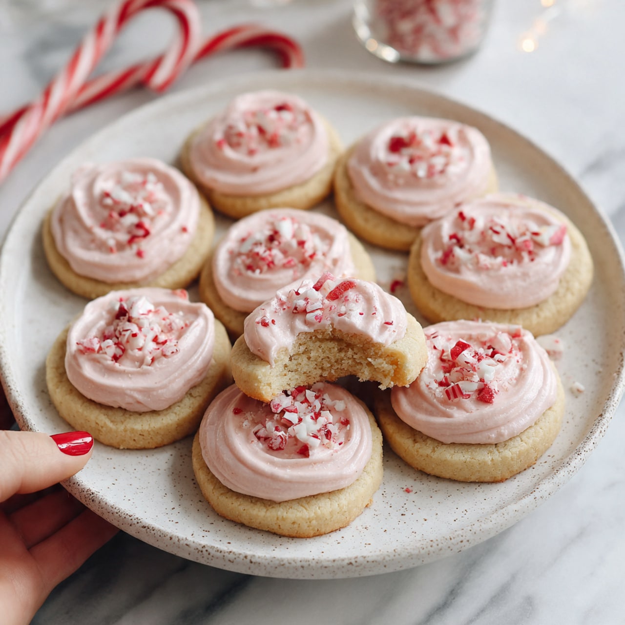 A white speckled plate holds eleven round shortbread cookies, each topped with one thick, swirled layer of pale pink frosting. The frosting has a smooth and creamy texture, spreading slightly beyond the cookie edges. On top of the frosting are small pieces of crushed red and white candy canes, scattered unevenly. One cookie is slightly raised above the others and has a bite showing a soft, crumbly light beige interior. The plate sits on a white marbled surface, with a woman's hand holding a candy cane in the background. Photo taken with an iphone --ar 4:5 --v 7