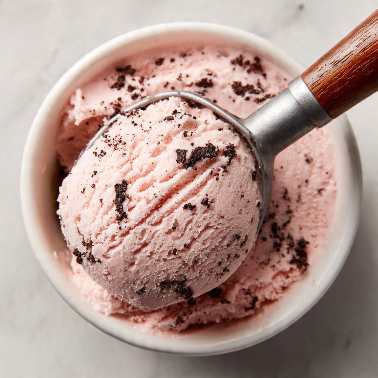 A close-up view of a single scoop of light pink ice cream with small dark bits mixed inside, held by an ice cream scooper with a light green handle and copper metal part, resting inside a white bowl filled with more of the same pink ice cream. The bowl is set on a white marbled surface. The texture of the ice cream is creamy and smooth with visible small chunks throughout. photo taken with an iphone --ar 4:5 --v 7