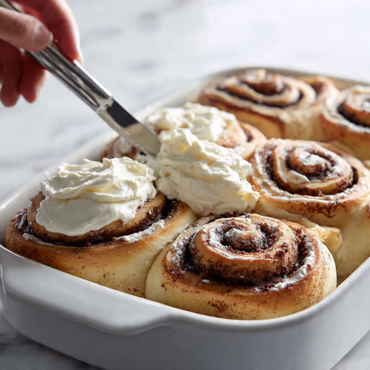 The image shows close-up of six cinnamon rolls in white baking pan arranged in two rows of three. Each roll has three visible swirled layers of light brown dough with darker brown cinnamon filling. On top of some rolls, a thick, creamy layer of white frosting is being spread with a metal spatula. The frosting has a smooth, swirled texture. The background surface is white marbled texture. photo taken with an iphone --ar 4:5 --v 7