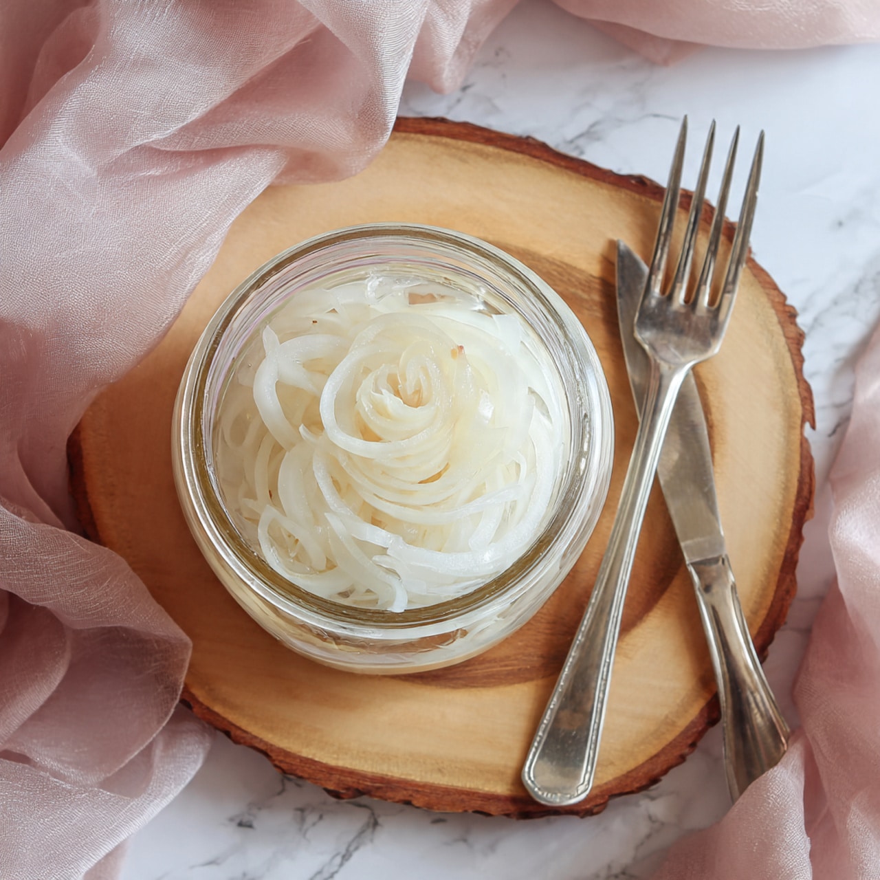 A clear glass jar filled with layers of thin, pale white onion slices soaked in a clear liquid, shown from above with the jar open and its white lid placed nearby on a wooden board with natural edges, set on a white marbled surface with a silver fork partially visible and soft pink fabric draped around the edges. photo taken with an iphone --ar 4:5 --v 7