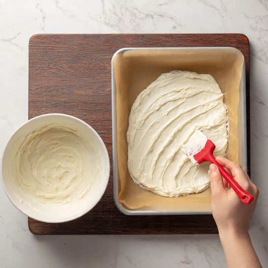 The image shows a baking tray lined with brown parchment paper filling most of the frame. On top, there is one layer of a light, creamy white spread being smoothed out by a red spatula held by a woman's hand from the right side. To the left of the tray, a white bowl contains more of the same creamy white mixture, with visible soft, creamy texture inside. The whole scene is set on a white marbled surface with a dark brown wooden background at the top edge. photo taken with an iphone --ar 4:5 --v 7