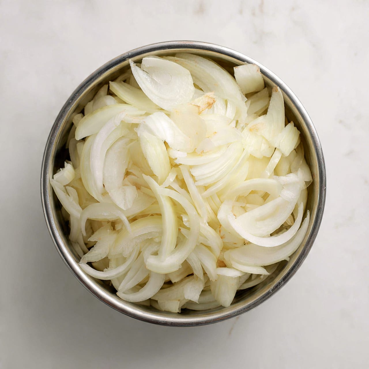 A metal bowl filled with many sliced white onions. The onion slices have thin and thick parts, some pieces are curved while others are straight, giving a mix of smooth and rough textures. The bowl sits on a white marbled surface, shining softly from the light above, highlighting the onions' white and pale yellow colors. The bowl is viewed from above, showing the layers of onion slices inside, piled loosely with no clear order. photo taken with an iphone --ar 4:5 --v 7