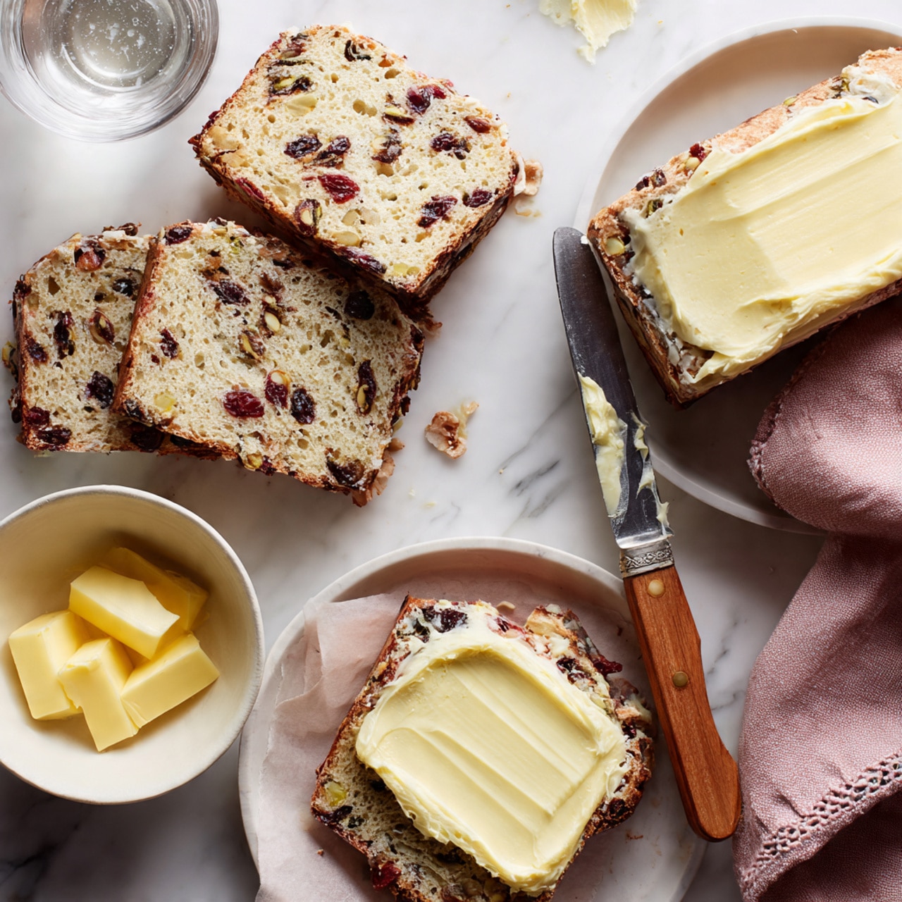 The image shows four slices of rustic fruit and nut bread with a coarse texture and visible pieces of dark red fruit and light brown nuts scattered throughout the light brown crumb. Two slices are placed on a white marbled surface near a butter knife with a wooden handle that has creamy yellow butter spread on its flat edge and some crumbs. Another slice with a thick layer of butter spread evenly covers most of its surface, resting on a white plate, with a fourth slice beside it. A small white bowl filled with soft golden yellow butter sits nearby, with a glass of water partially visible at the top left. A folded dusty pink napkin with a stitched edge is on the right side of the frame. The background is a white marbled texture photo taken with an iphone --ar 4:5 --v 7