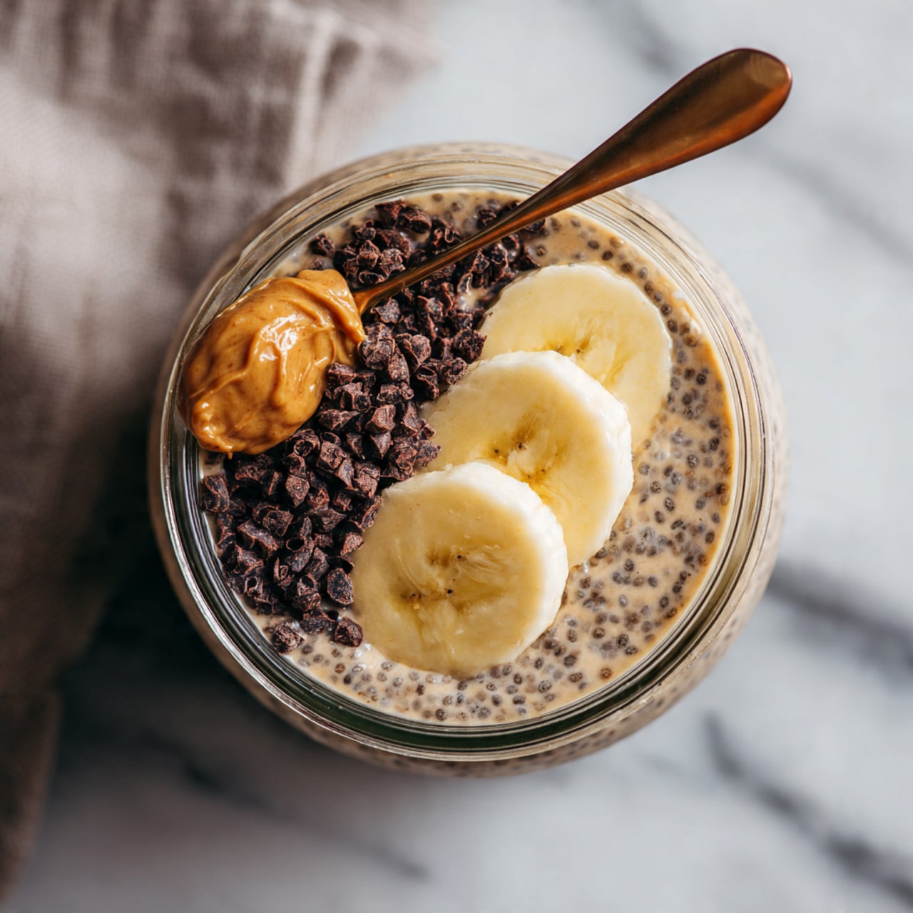 A clear jar filled with a light brown, creamy chia pudding base with visible chia seeds throughout. On top, there are three round slices of fresh banana arranged in a row, a spoonful of thick, golden almond butter placed next to the banana slices, and a small pile of dark brown cacao nibs on the side. The jar is placed on a white marbled surface with a blurred cloth in the background. Photo taken with an iphone --ar 4:5 --v 7