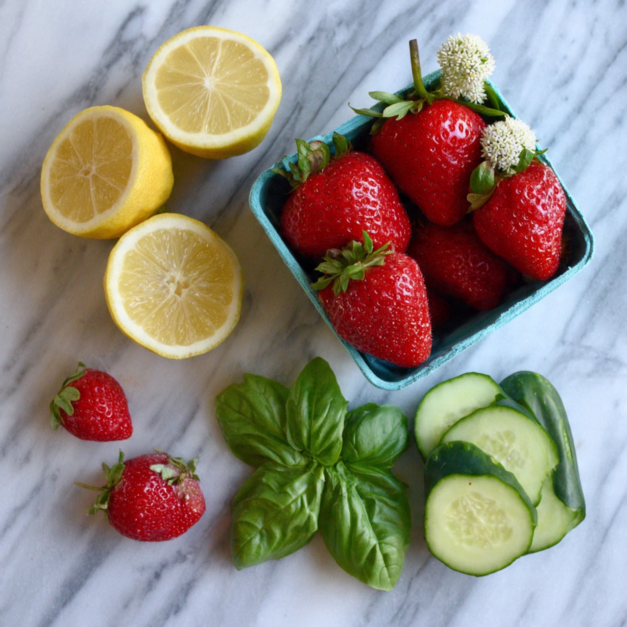 The image shows a small blue container filled with fresh, red strawberries with green stems and a few white flowers on top, sitting near the top right corner. To the left, there are five lemon halves, showing their juicy, pale yellow inside. Below the lemons, there are a few loose strawberry leaves and a couple of small strawberries with stems attached. Near the bottom right, there are three light green cucumber slices, next to a bundle of fresh green basil leaves. All items are arranged on a white marbled surface. photo taken with an iphone --ar 4:5 --v 7