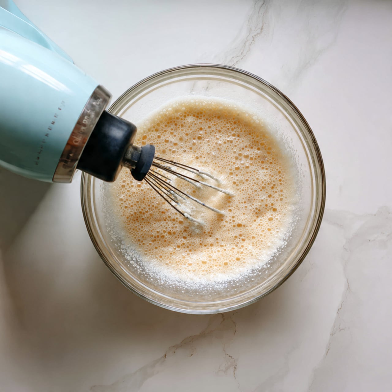 A close-up of a single brown soft bread roll with a shiny topside sprinkled with light beige oat flakes, sitting on a white plate with a simple ridged edge. The roll is fluffy and torn slightly on one side, showing a light tan inner texture. In the background, a white marbled surface holds a clear glass baking dish lined with brown parchment paper filled with more similar bread rolls. To the side, there is a partial view of a small bowl with a light yellow spread. photo taken with an iphone --ar 4:5 --v 7