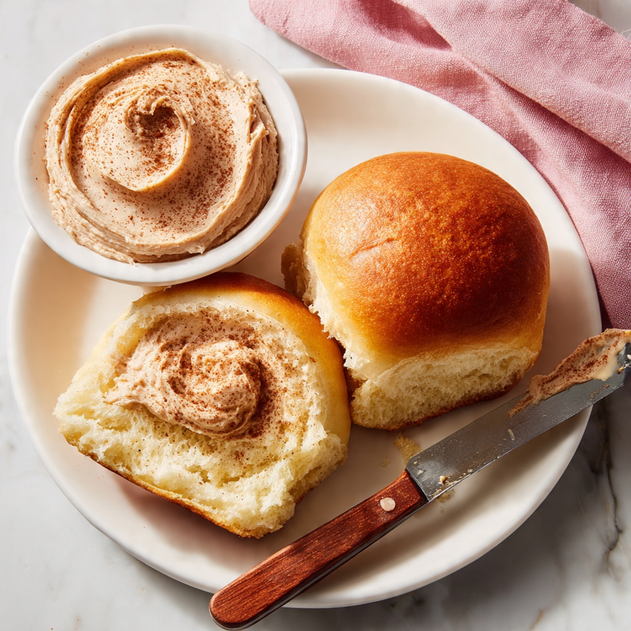 A glass baking dish holds nine golden brown, soft dinner rolls arranged in three rows and three columns on a white marbled surface. The rolls have smooth tops with a shiny finish and a light sprinkling of coarse salt on some. One roll near the corner is partially pulled apart, revealing its fluffy, airy inside with a spread of light brown cinnamon butter melting into the soft dough. Next to the dish, a butter knife with a light wooden handle and gold blade rests on the surface, covered in swirls of the cinnamon butter. photo taken with an iphone --ar 4:5 --v 7
