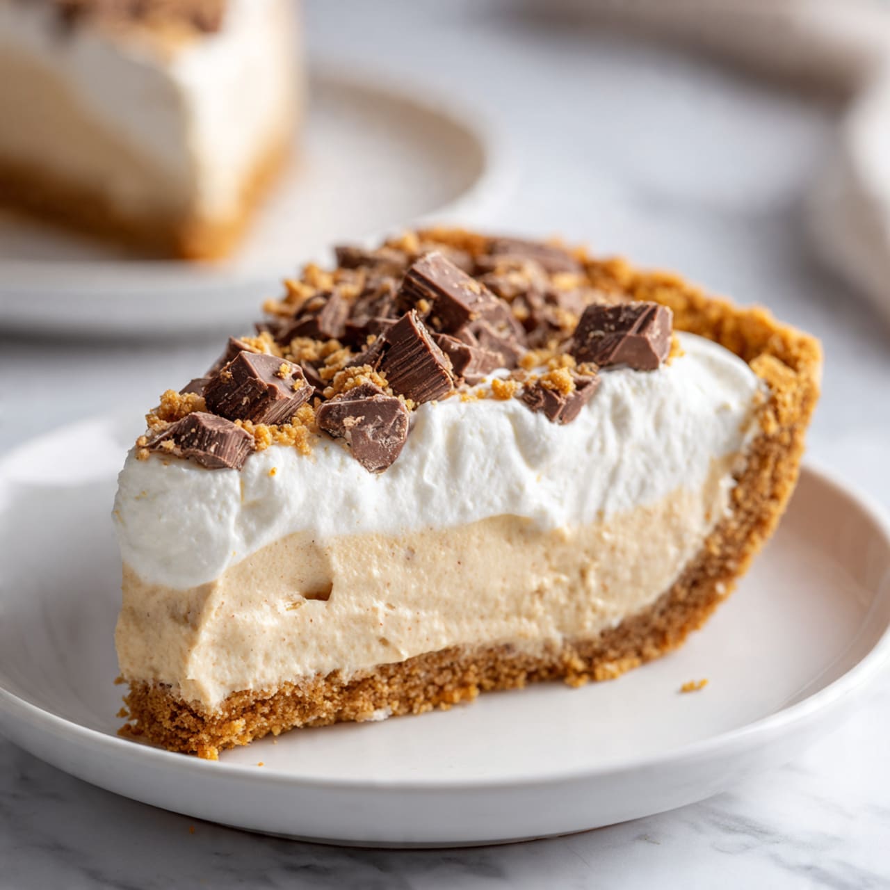 A slice of pie on a white plate sits on a white marbled surface. The pie has three visible layers: a golden-brown crumbly crust at the bottom and sides, a thick, creamy light tan filling in the middle with a smooth and slightly airy texture, and a fluffy white whipped topping covering the filling. On top of the whipped layer, there are scattered chunks of chocolate pieces, adding texture and color contrast. In the background, another slice of pie on a white plate is slightly out of focus. Photo taken with an iphone --ar 4:5 --v 7