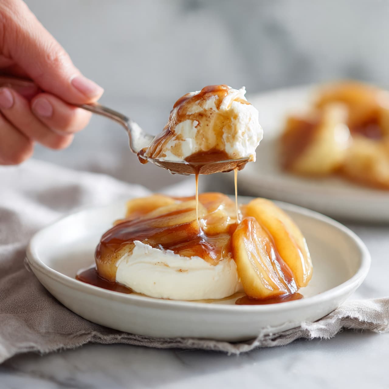 The image shows a clear glass filled with layers of white ice cream and light brown caramel sauce. The glass has three visible layers, starting with ice cream at the bottom, followed by caramel sauce, and then ice cream again. On top, there are a few small chunks of cooked apple pieces covered in caramel that drip down the side of the glass. A bronze spoon is placed inside the glass from the left side. The background is a white marbled surface with a blurred plate of caramel and apple pieces in the back right. Photo taken with an iphone --ar 4:5 --v 7