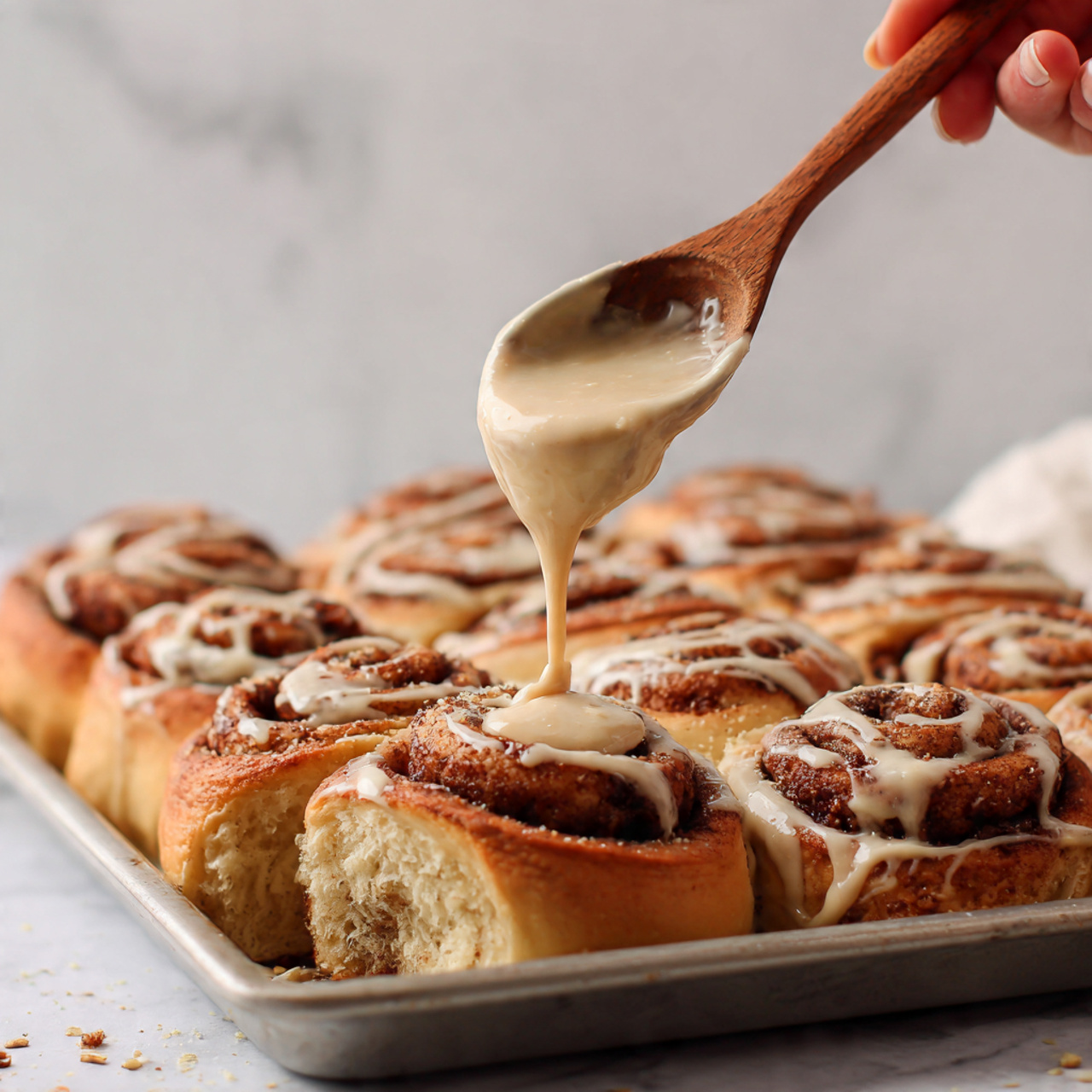 The image shows a close-up of nine cinnamon rolls in a white baking dish, arranged in a 3x3 grid. Each roll has golden-brown swirled layers with a darker cinnamon filling creating a spiral pattern. A thick, creamy beige frosting is being spread on the upper middle rolls with a wooden spatula held by a woman's hand, covering part of the swirls. The cinnamon rolls have a soft, fluffy texture with slightly crisp edges. The background is a white marbled surface. photo taken with an iphone --ar 4:5 --v 7