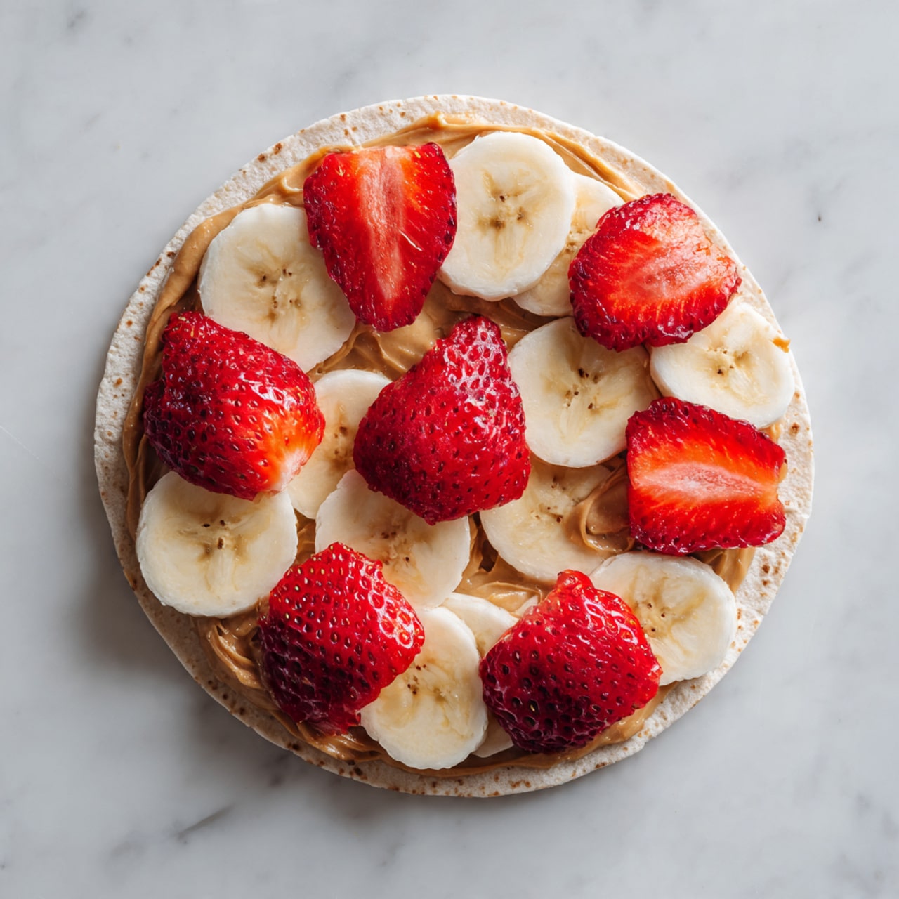 A round tortilla lays flat on a white marbled surface, spread evenly with a thick layer of light brown peanut butter that reaches near the edges. On top of the peanut butter, there is a layer of thin, curved banana slices arranged side by side, mostly covered by a final layer of bright red strawberry slices, which are oval and slightly translucent, showing the berry's seeds and texture. The strawberry pieces are spread out evenly, fully covering the bananas underneath. Photo taken with an iphone --ar 4:5 --v 7