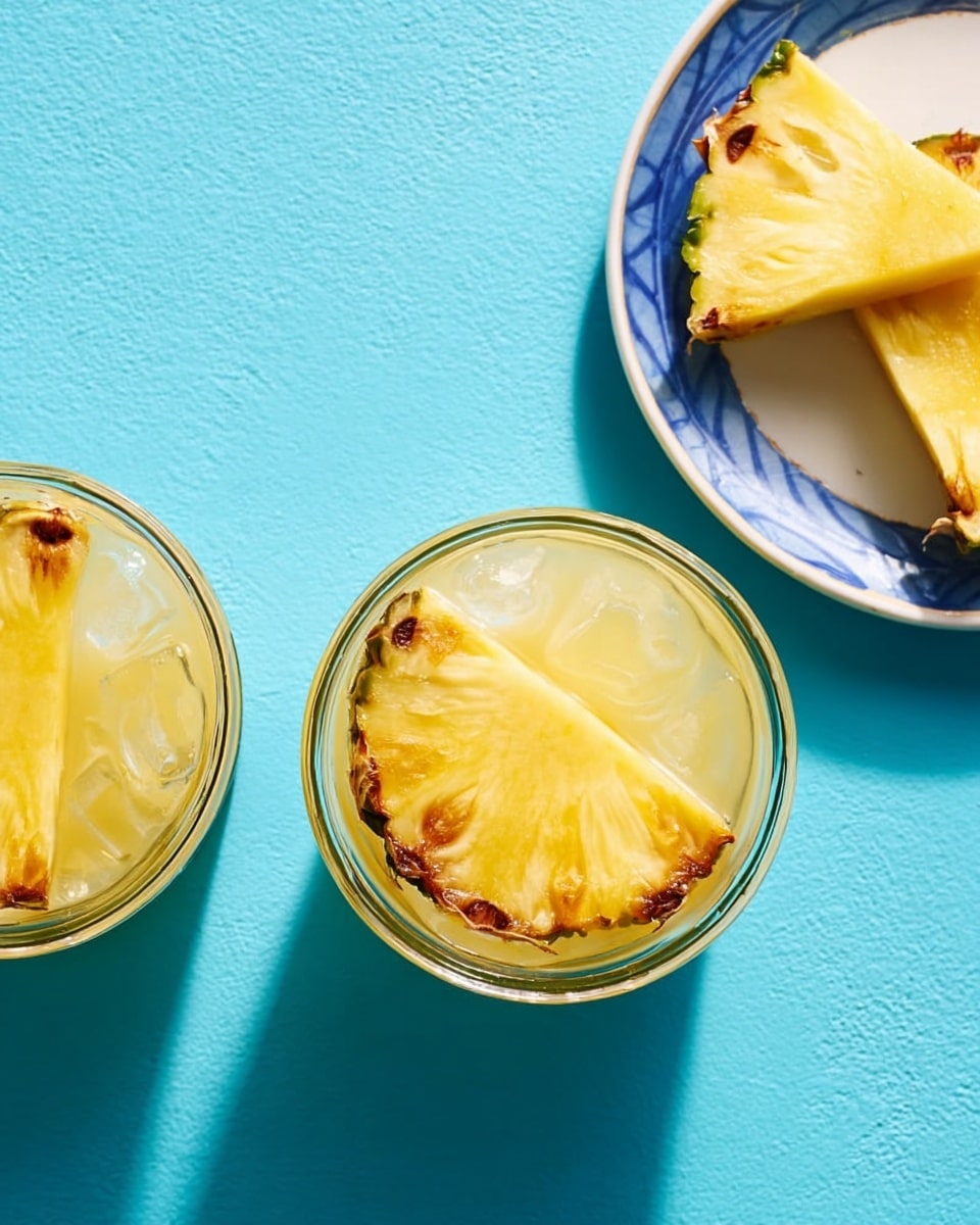 Two short clear glasses filled with light creamy yellow frozen drinks showing swirled icy texture. Each drink is topped with a single thick slice of pineapple ring placed inside the glass rim, with bright green pineapple leaves standing tall behind the ring. The glasses sit on a smooth light blue surface with soft shadows. photo taken with an iphone --ar 4:5 --v 7