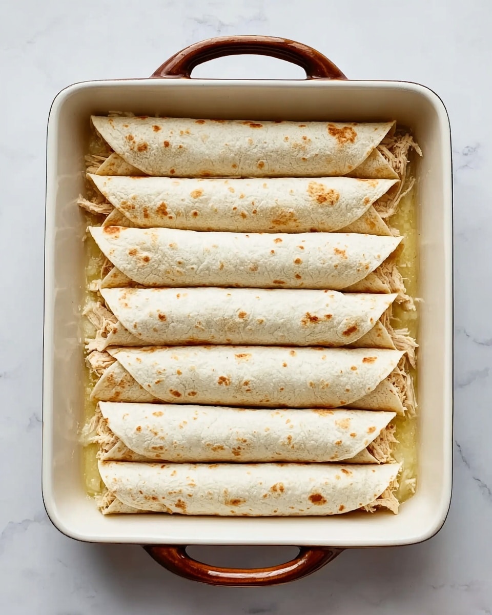 This dish shows two rolled pieces of enchiladas placed side by side inside a white rectangular pan with brown handles. Each roll is covered with a thick layer of melted white cheese sprinkled with small green herb bits and tiny red flakes. The cheese stretches slightly between the two enchiladas, revealing some melted texture beneath. The bottom layer consists of a light green sauce with a slightly oily texture pooling around the base of the enchiladas. Fresh parsley leaves are placed on the white marbled surface near the pan. Photo taken with an iphone --ar 4:5 --v 7