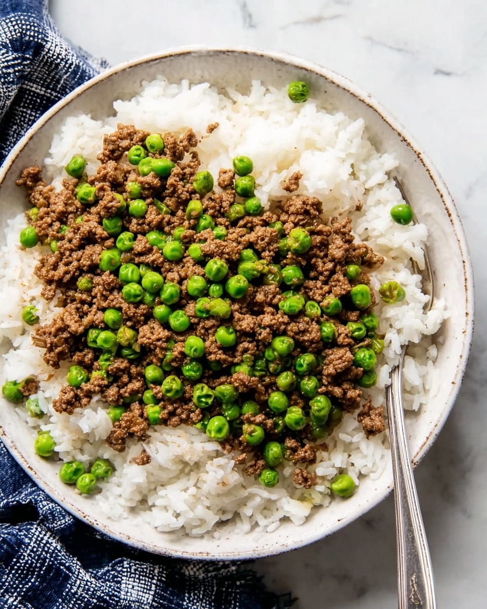 A white bowl filled with a base layer of fluffy white rice, topped with a layer of cooked ground meat mixed with bright green peas scattered evenly over the surface. The rice grains look soft and separate, while the meat and peas add a textured and colorful contrast on top. A silver spoon rests inside the bowl on the right side, partially buried in the food. The bowl is placed on a white marbled surface with a dark blue and white checked cloth next to it. photo taken with an iphone --ar 4:5 --v 7
