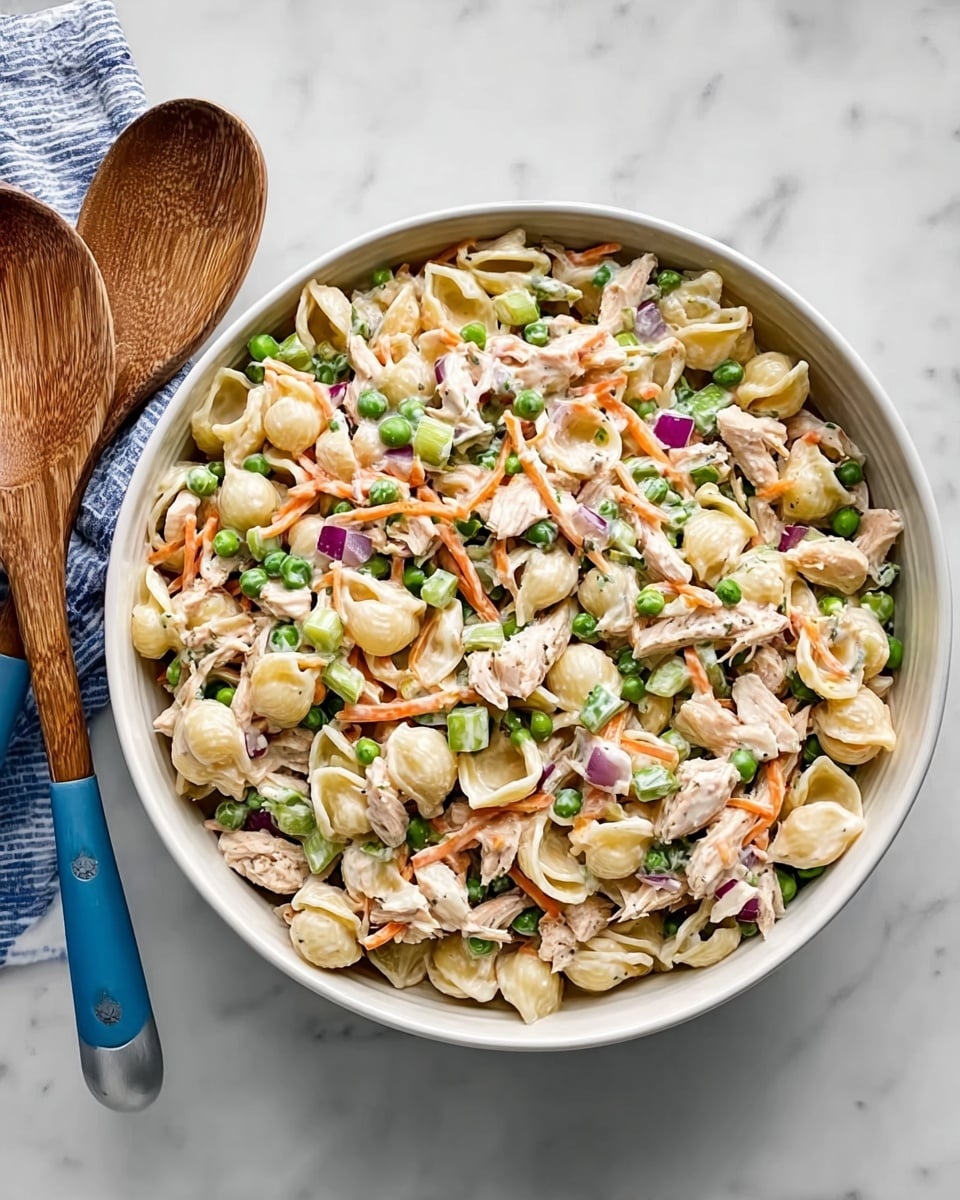 A large white bowl sits on a white marbled surface, filled with a pasta salad made of shell-shaped pasta mixed with shredded light beige chicken pieces, bright green peas, thin orange carrot strips, small diced purple onions, and pale green celery chunks. The salad is coated lightly with a creamy white dressing that blends all the ingredients together. Two wooden serving utensils with blue painted handles rest next to the bowl. photo taken with an iphone --ar 4:5 --v 7