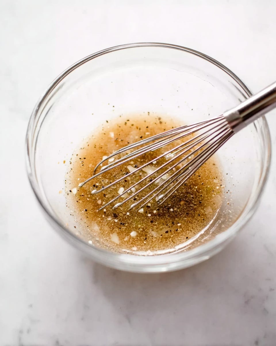A clear glass bowl sits on a white marbled surface, filled with a light brown liquid mixture with small bits of white and black, likely finely chopped onions and black pepper. A silver whisk rests inside the bowl, partially submerged in the mixture. The overall look is fresh and simple, with the bowl and whisk casting soft shadows on the bright white marbled background. photo taken with an iphone --ar 4:5 --v 7