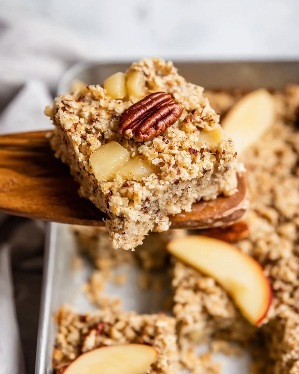 The image shows a close-up of a square oat bar with visible small pieces of pecans mixed throughout the top and sides, giving it a crumbly texture. Inside the bar, chunks of light yellow apples can be seen, adding contrast to the oatmeal color. The bar is held by a wooden spatula above a metal pan filled with more of the oat bars and some apple slices, all placed on a white marbled surface. The colors are mainly light beige, brown from the nuts, and pale yellow from the apple pieces. photo taken with an iphone --ar 4:5 --v 7