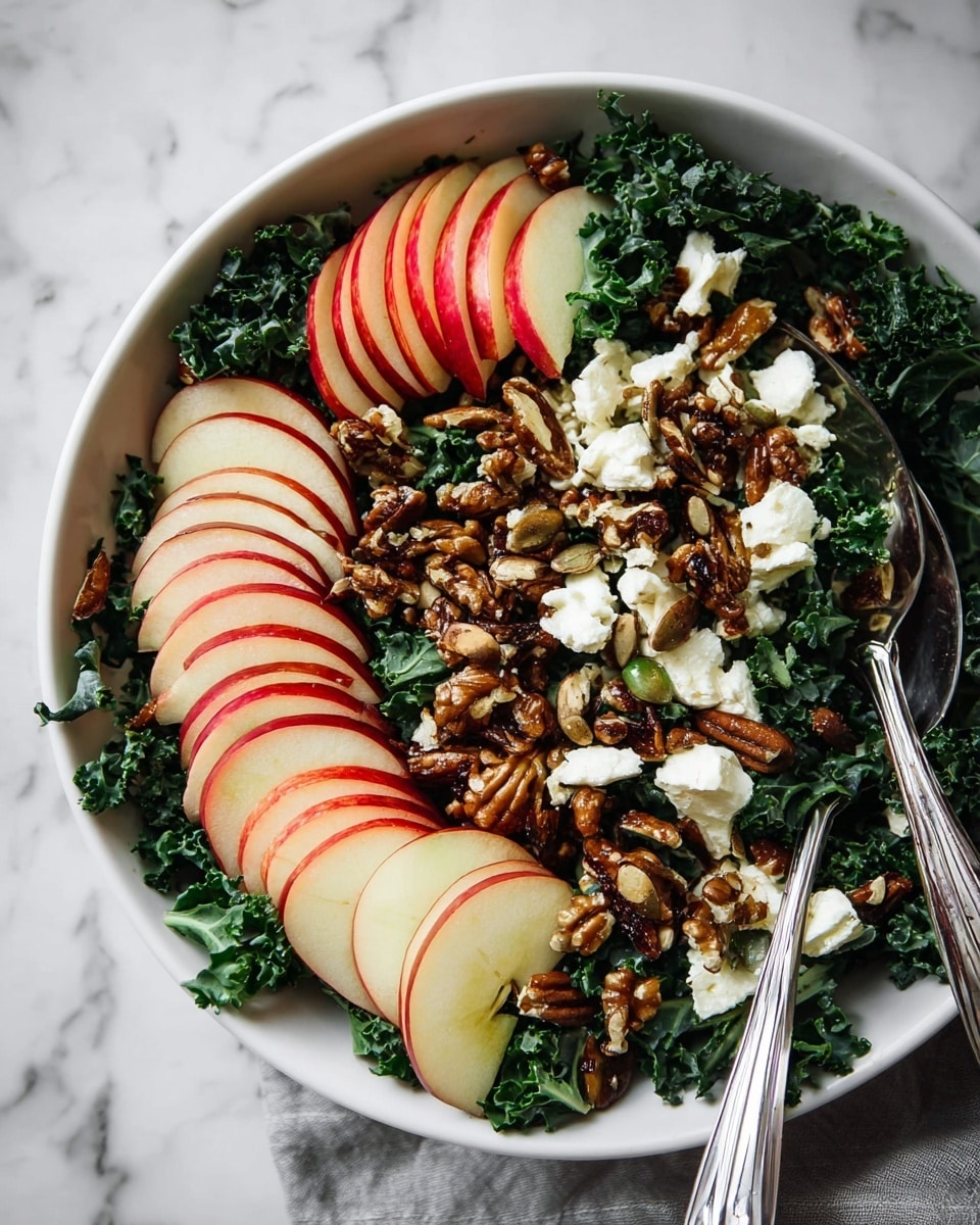 A white bowl filled with three main layers: the bottom layer is dark green curly kale leaves; the middle layer shows thinly sliced red and white apple pieces arranged in small stacks around the bowl; the top layer contains clusters of brown toasted seeds and nuts, along with small dollops of white soft cheese scattered evenly over the salad. Two silver spoons rest on the side inside the bowl, and the background shows a white marbled surface. photo taken with an iphone --ar 4:5 --v 7