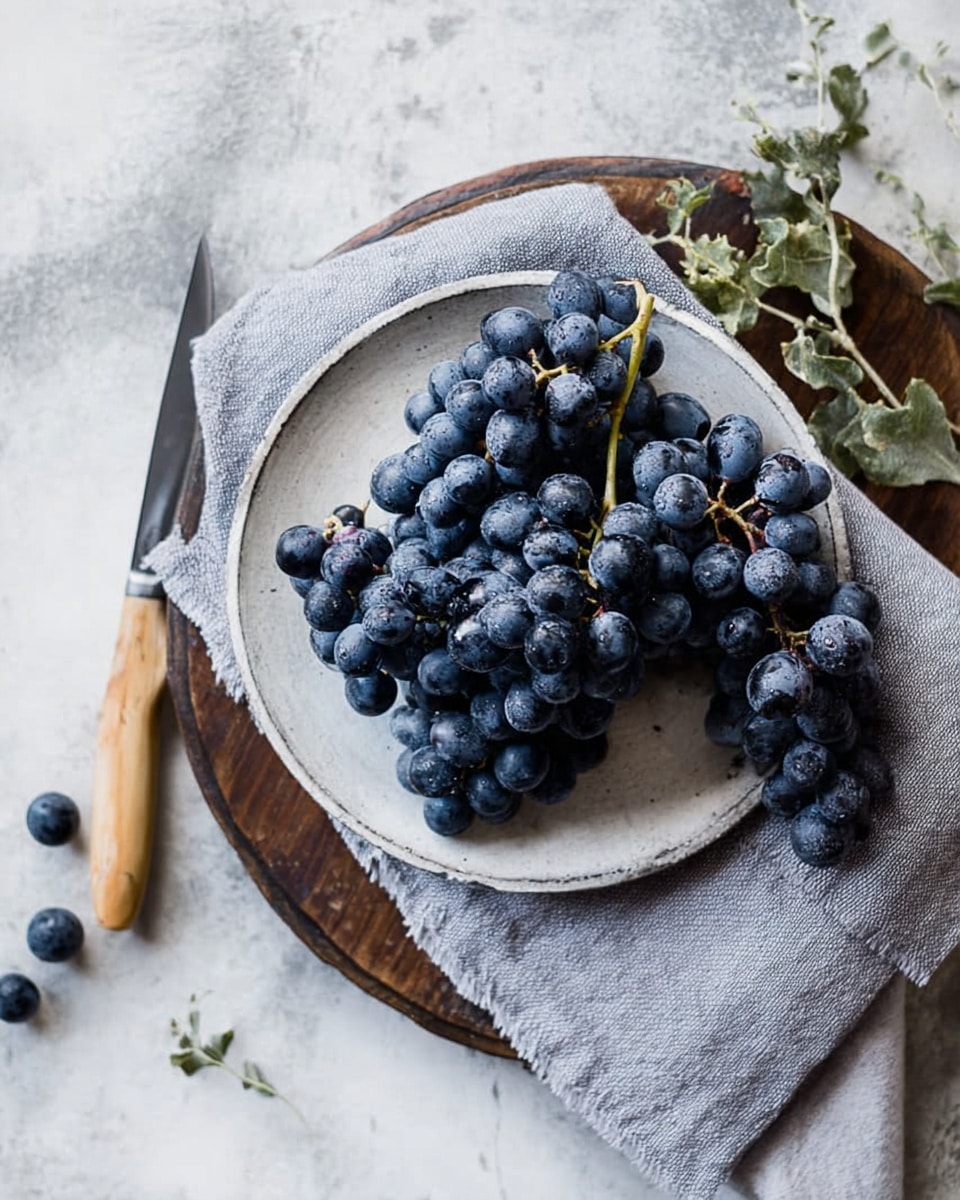 A bunch of dark blue grapes is placed on a small white round plate with a slightly rough texture, which sits on top of a folded light gray cloth. The plate and cloth rest on a round wooden board with a knife with a light wooden handle lying next to the grapes. Some small green leaves are scattered around the board and cloth. The entire setup is on a white marbled surface. photo taken with an iphone --ar 4:5 --v 7