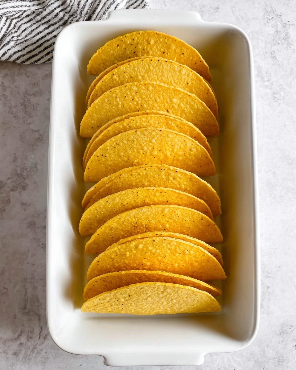 A white dish holds multiple tacos arranged upright, each taco has three layers: at the bottom, a crispy yellow corn shell; in the middle, a layer of cooked ground meat with a rich brown texture; on top, layers of bright green guacamole, white sour cream dollops, pink pickled onion slices, red diced tomatoes, and finely chopped green cilantro. There is shredded pale green lettuce peeking from under the toppings. A lime wedge is placed near the front tacos. The dish sits on a white marbled surface, and in the blurred background, small white bowls hold guacamole and red salsa. Photo taken with an iphone --ar 4:5 --v 7