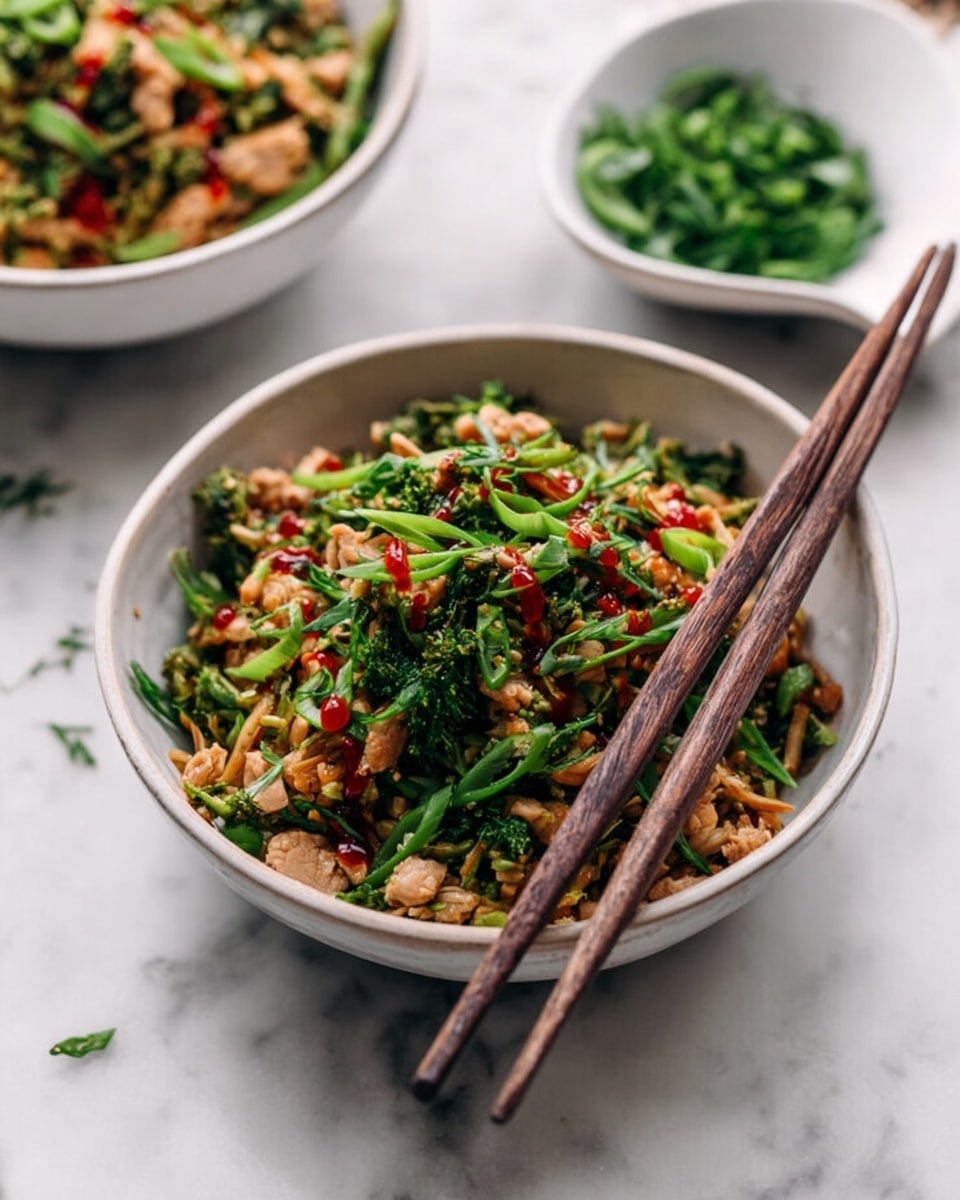 A white bowl filled with a mix of shredded light brown and green vegetables as the base layer, topped with small pieces of cooked light tan meat scattered throughout. Bright green chopped herbs and thin green onion slices are spread richly on top, with a few drops of dark red sauce drizzled over the dish. Two dark brown wooden chopsticks rest across the rim of the bowl. The bowl is set on a white marbled surface, with another similar bowl slightly blurred in the background and a small white dish of chopped green herbs partially shown nearby. Photo taken with an iphone --ar 4:5 --v 7