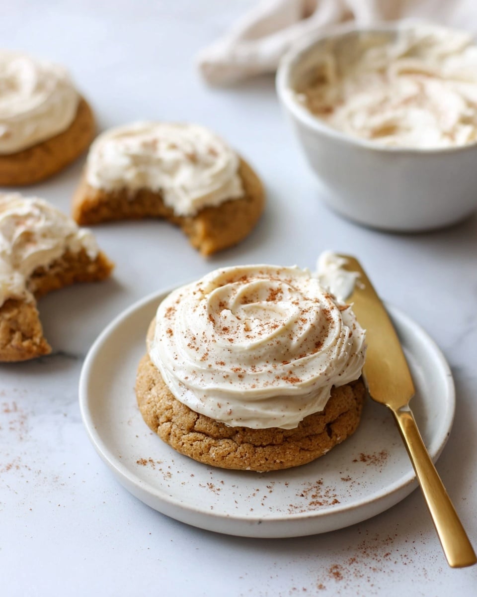 The image shows soft round cookies on a white marbled surface. Each cookie is golden brown with a rough texture and has a thick swirl of creamy white frosting on top. There are light sprinkles of brown spice powder over the frosting and some scattered on the surface. In the background, a white plate holds more cookies with a butter knife lying across it, with some frosting on the knife. A dark cloth is beneath the plate. The scene is bright and simple, focusing on the cookies’ details. Photo taken with an iphone --ar 4:5 --v 7