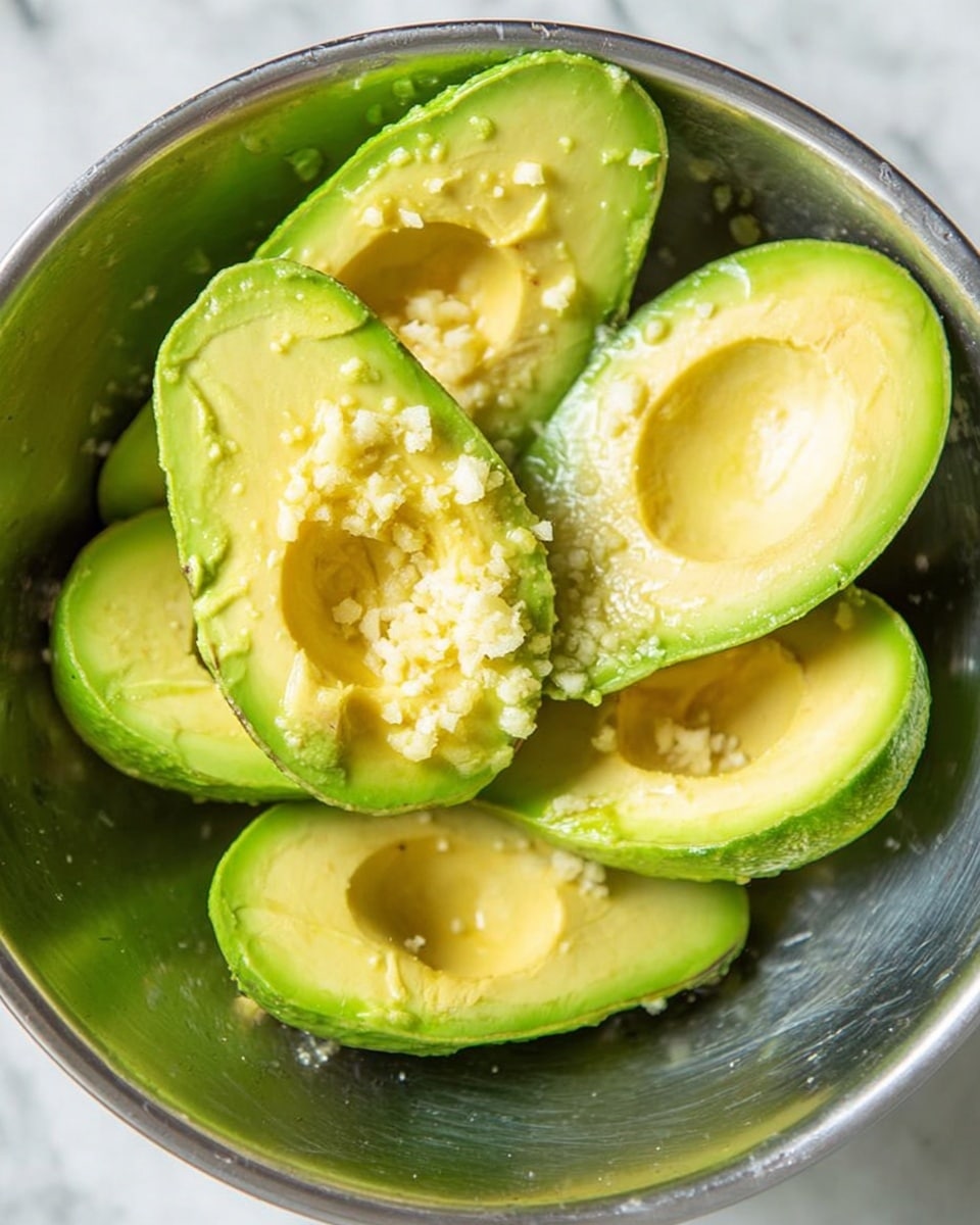 The image shows a close-up of a food bowl layered with white rice at the bottom, topped with pieces of grilled chicken that are golden brown with char marks, a colorful mix of yellow corn and red bell pepper bits, and a dollop of bright green mashed avocado on top. Next to this bowl is a wooden bowl filled with more mashed avocado that has a creamy, slightly chunky texture, with a wooden spoon inside. In the background, there is a halved avocado with its seed visible and halved limes on a white marbled surface. Photo taken with an iphone --ar 4:5 --v 7