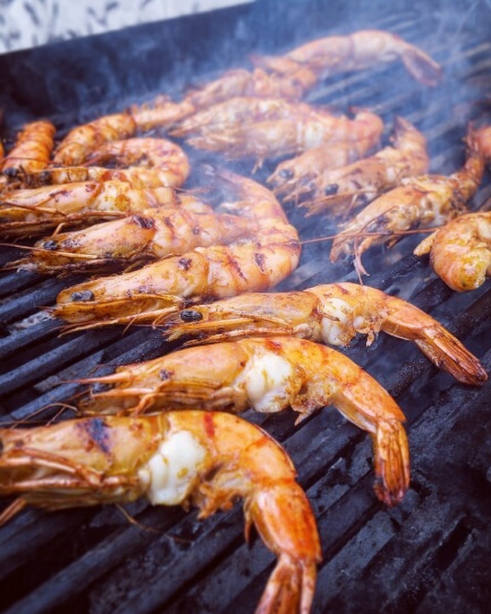 The image shows several grilled shrimp skewers arranged closely on a white plate, each shrimp having a shiny, orange-brown color with slight char marks. The shrimp are garnished with small pieces of purple-red onion and green parsley scattered on top. Lime wedges with light green flesh appear between the shrimp on the skewers, adding a fresh contrast. The background is a white marbled surface. photo taken with an iphone --ar 4:5 --v 7