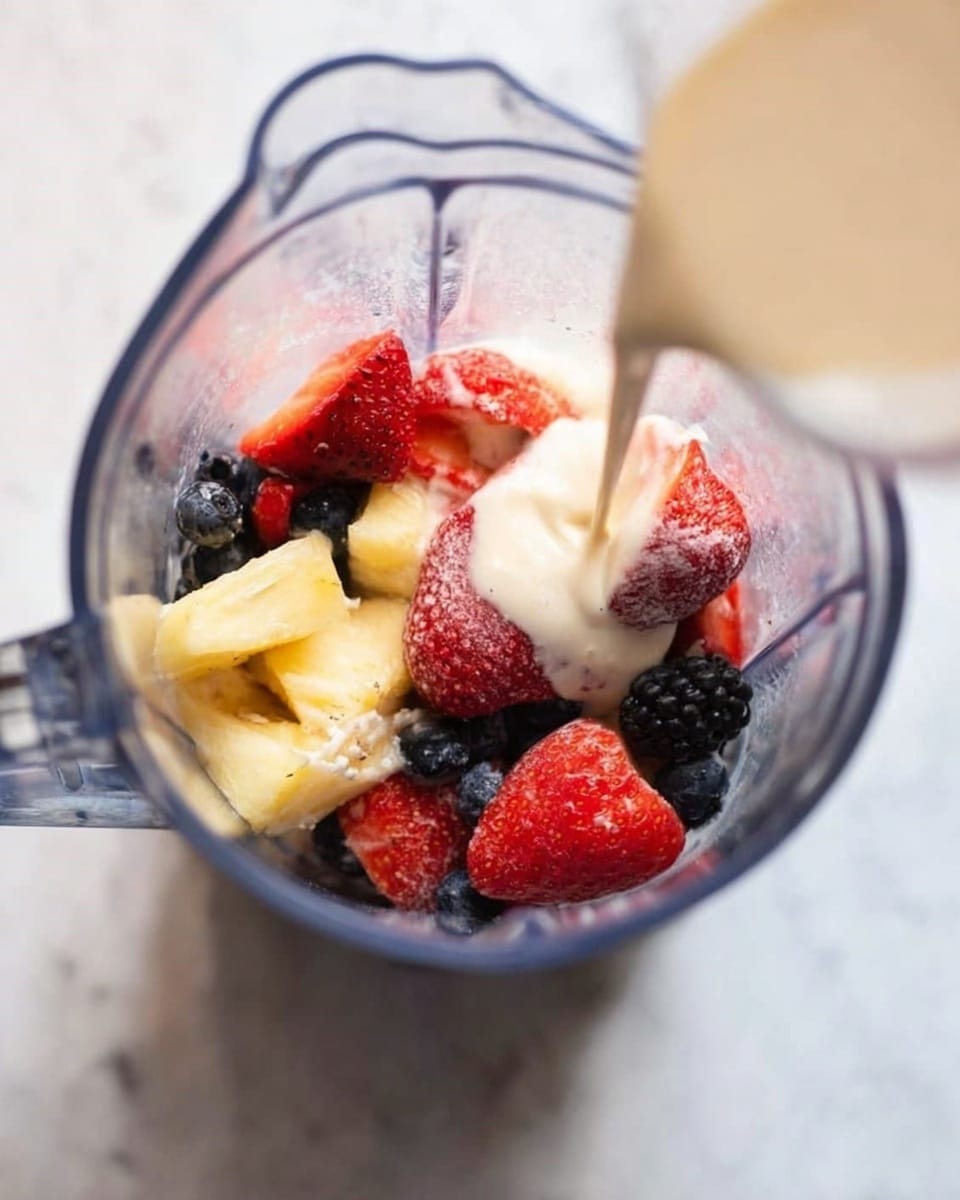 A tall clear glass jar filled with a thick light pink smoothie that has small dark specks inside, topped with three fresh whole blueberries placed on the smooth surface. Around the jar, several loose blueberries are scattered on a white marbled surface, and a small white bowl filled with more blueberries is blurred in the background. The overall look is fresh and inviting, focusing on the creamy texture and vibrant colors of the smoothie and berries. photo taken with an iphone --ar 4:5 --v 7