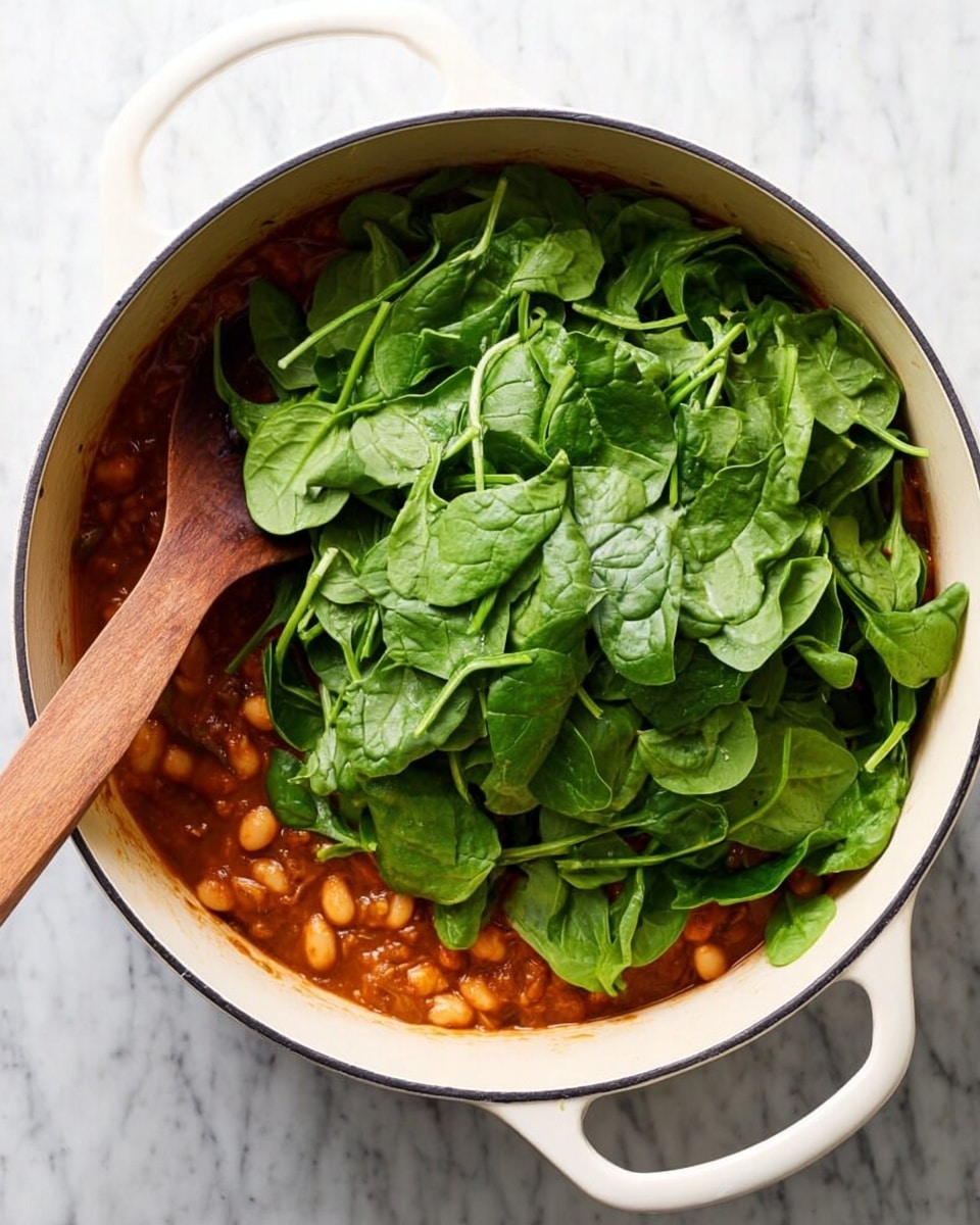 A white round plate holds a dish with three main layers: the base layer is soft, round gnocchi in a light orange creamy sauce mixed with wilted dark green spinach and slices of brown mushrooms; the middle layer shows the sauce coating the gnocchi and mushrooms, creating a slightly shiny texture; the top layer is scattered with thin, irregular white cheese shavings and small fresh green basil leaves. A gold spoon rests inside the plate, leaning against the food. The plate is placed on a striped cloth on a white marbled surface with some herbs around. Photo taken with an iphone --ar 4:5 --v 7
