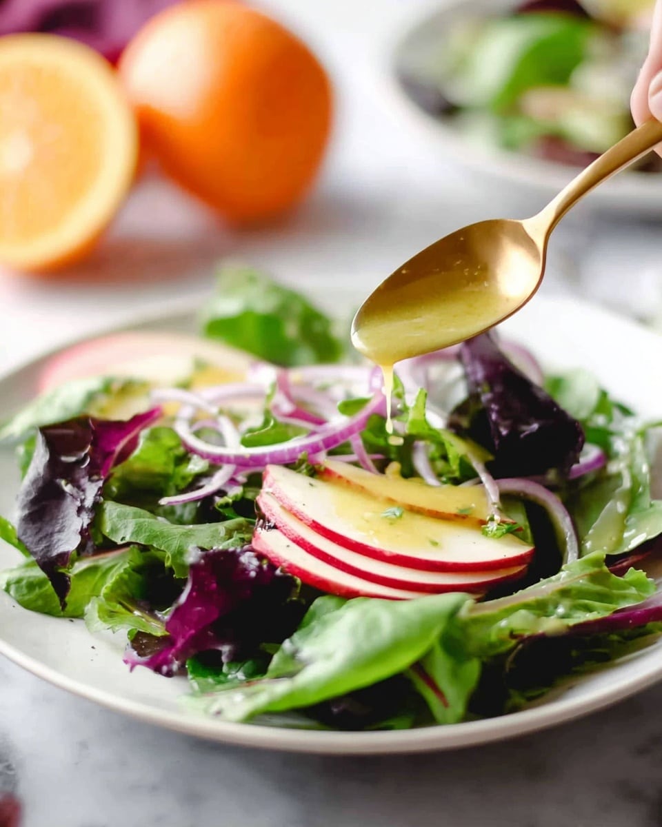 A white plate on a white marbled surface holds a fresh salad with two layers: the base layer is made up of mixed green and purple leafy greens with smooth and crinkled textures, and the top layer features thin slices of red apple and rings of purple onion scattered evenly. A woman's hand holding a gold spoon drizzles a creamy yellow dressing over the salad, adding a glossy finish. In the background, a halved orange and a second plate of salad are softly blurred. photo taken with an iphone --ar 4:5 --v 7