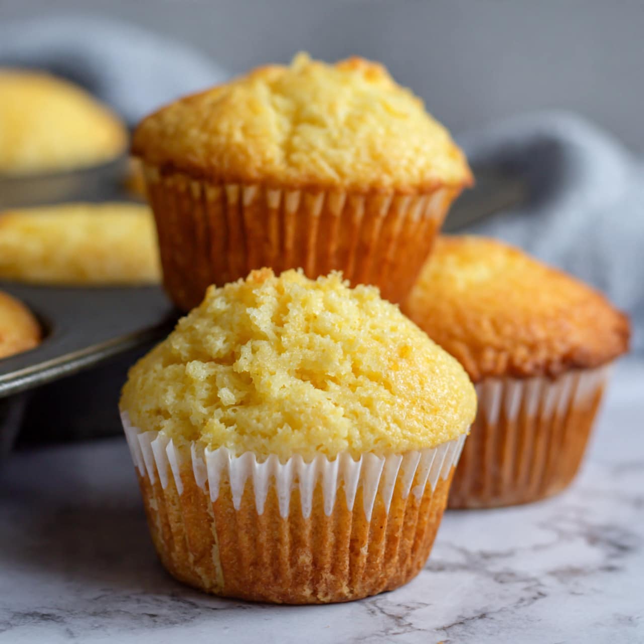 A close-up view of two golden yellow muffins in white paper liners, set in a dark muffin tray on a white marbled surface. The muffins have a smooth, slightly domed top with a soft texture. One muffin is upright, showing the full height with ridged liner details, while the other is lying flat, displaying its rounded top and edge. The background is softly blurred with faint white and warm tones. Photo taken with an iphone --ar 4:5 --v 7